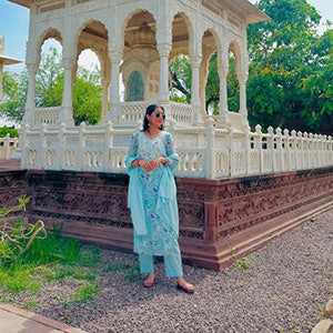 Woman in a light blue traditional outfit standing in front of a white architectural structure with arched windows.