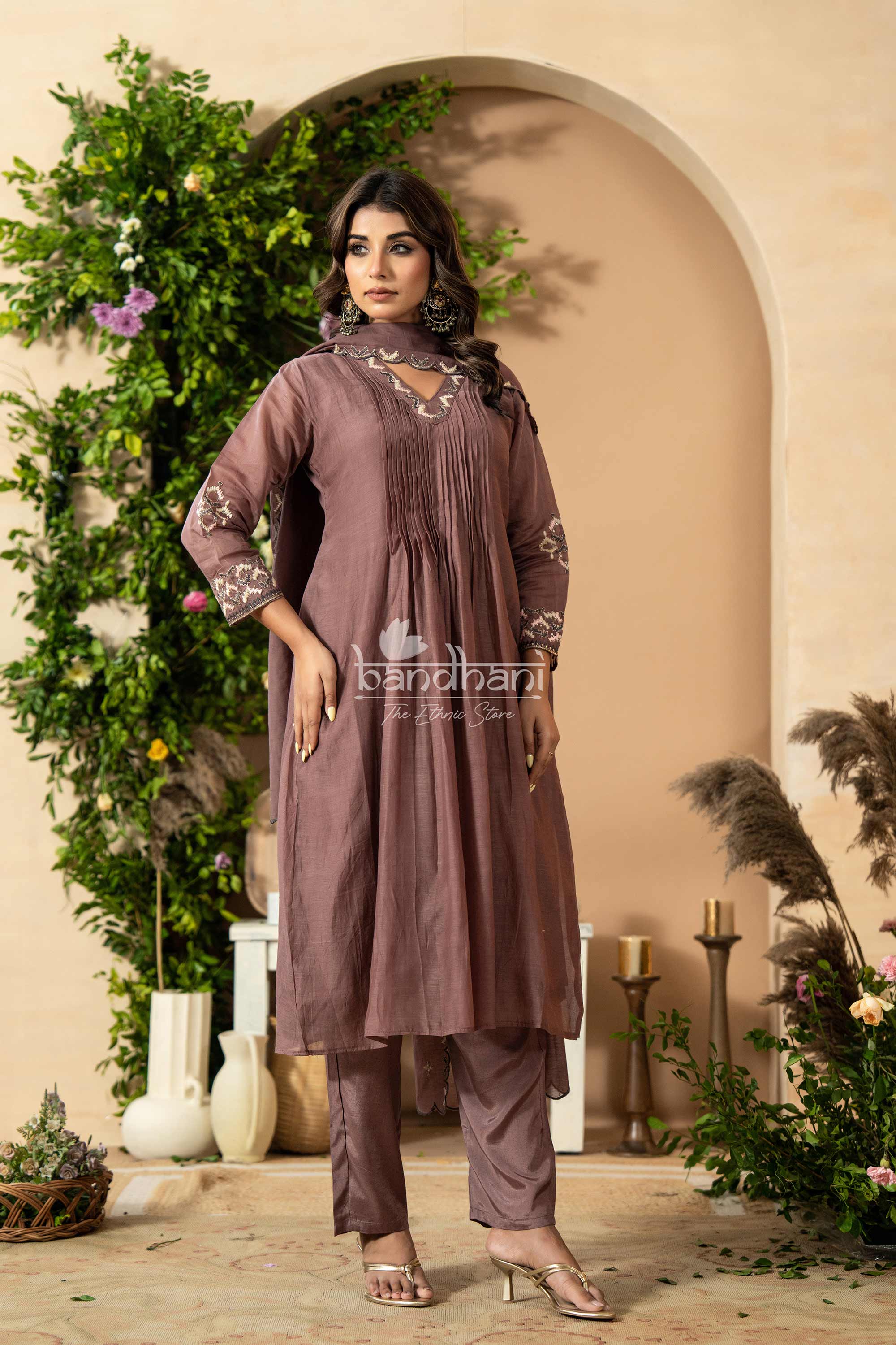 Woman wearing a brown traditional outfit with floral embroidery in a decorated indoor setting.