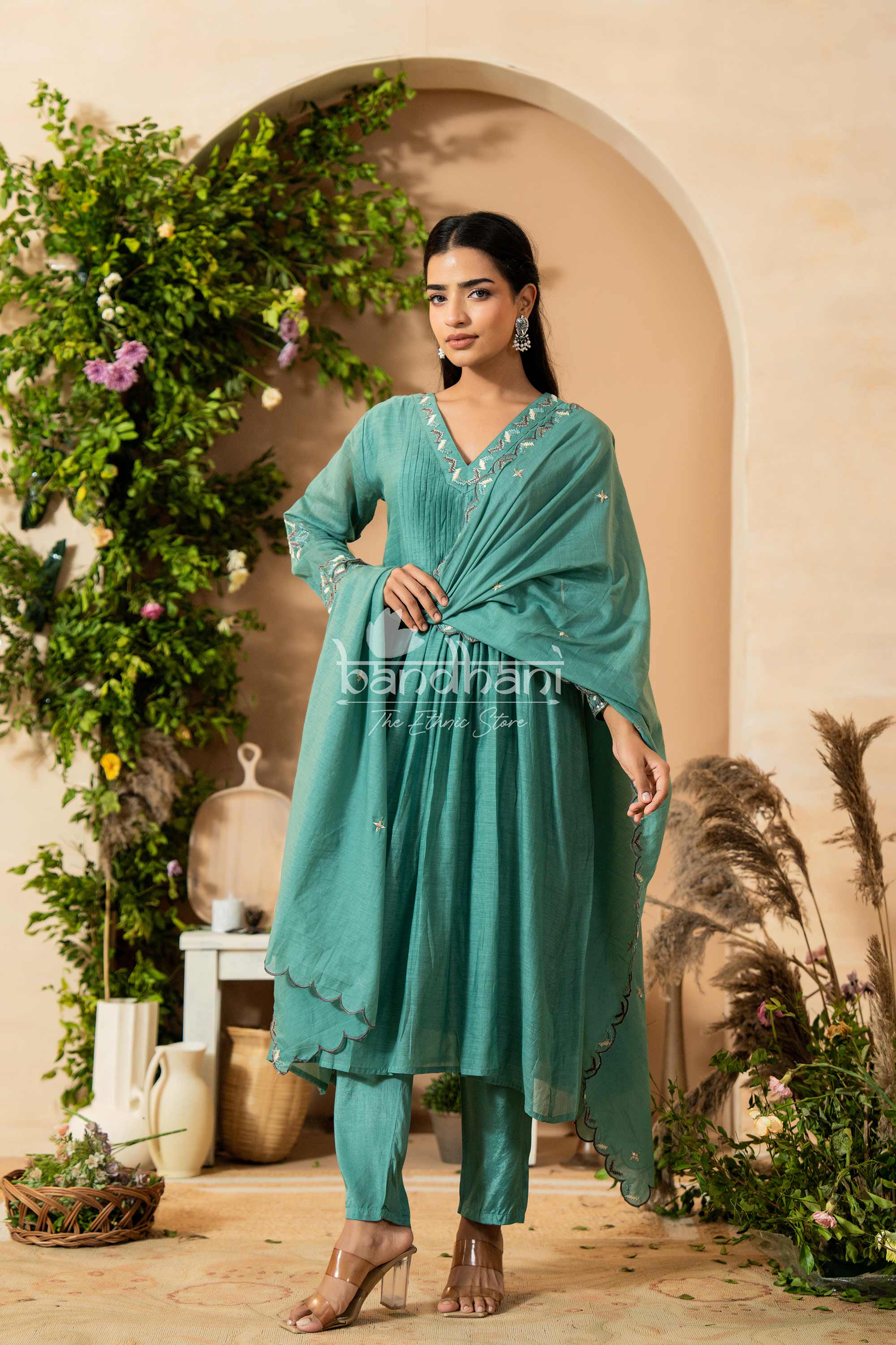 Woman in a teal traditional outfit standing in a decorative setting with plants.