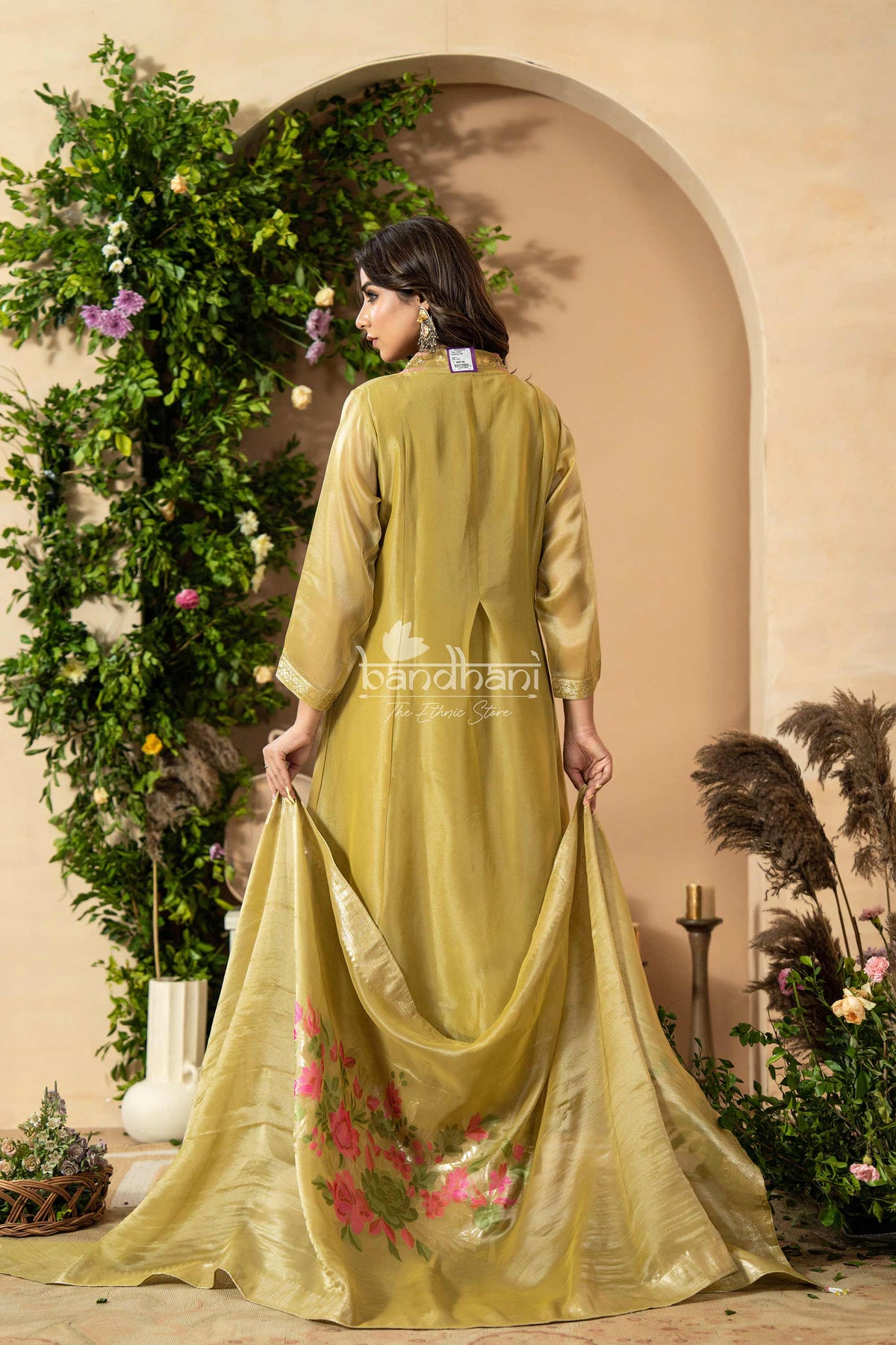 Woman in a green traditional outfit with floral design, standing in front of a decorative archway with plants.