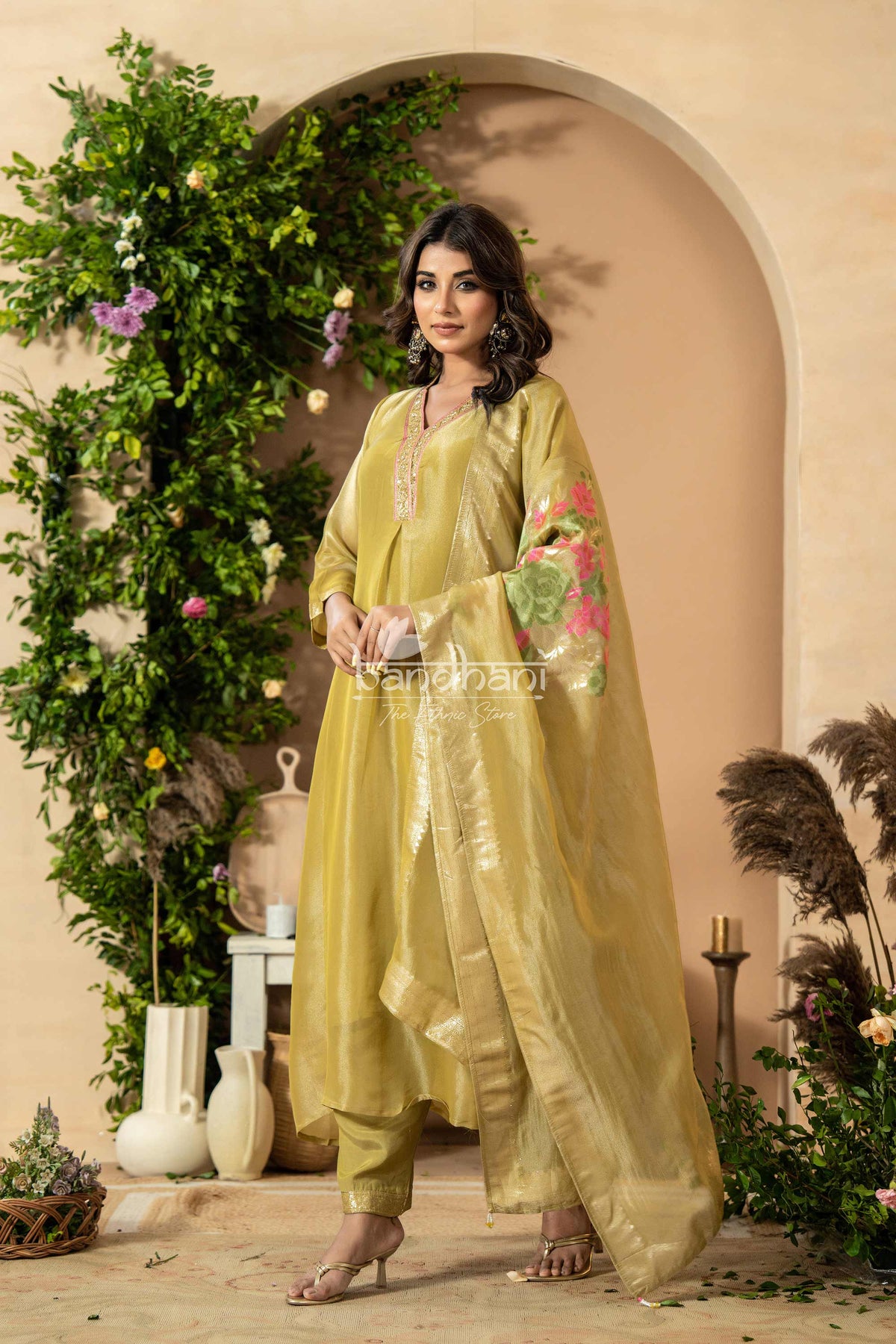 Woman in a light green traditional outfit standing in a decorated indoor setting with plants and flowers.