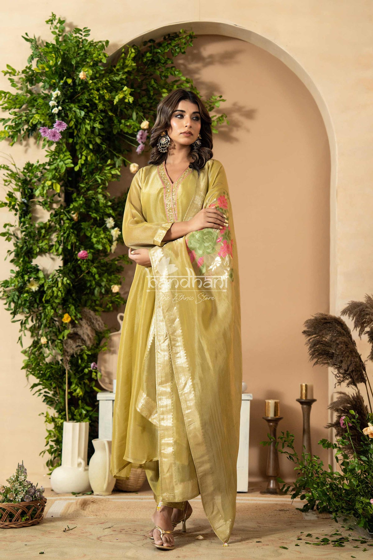 Woman in a light green traditional outfit standing in a decorated room with plants and candles.