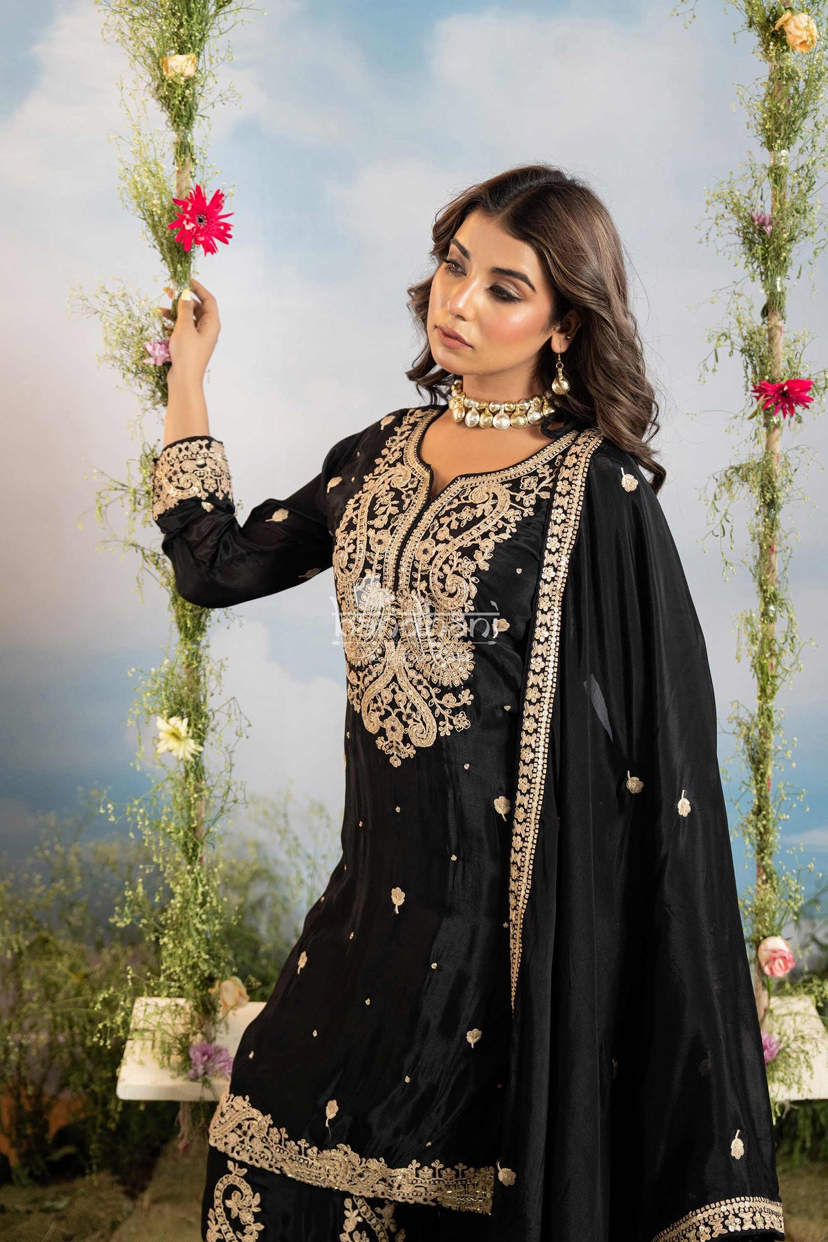 Woman in a black and gold embroidered traditional outfit with floral decorations in the background