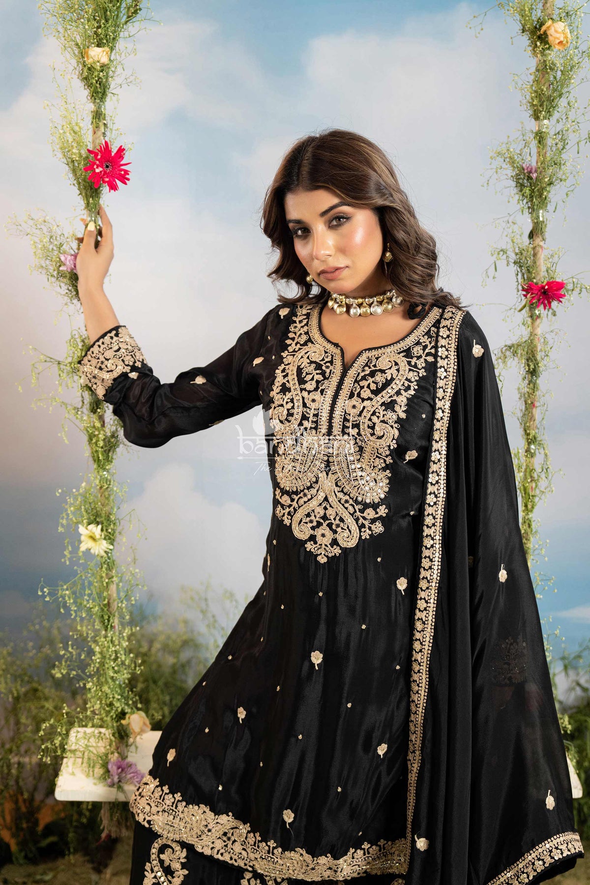 Woman in a black and gold embroidered traditional outfit standing outdoors with floral decorations.