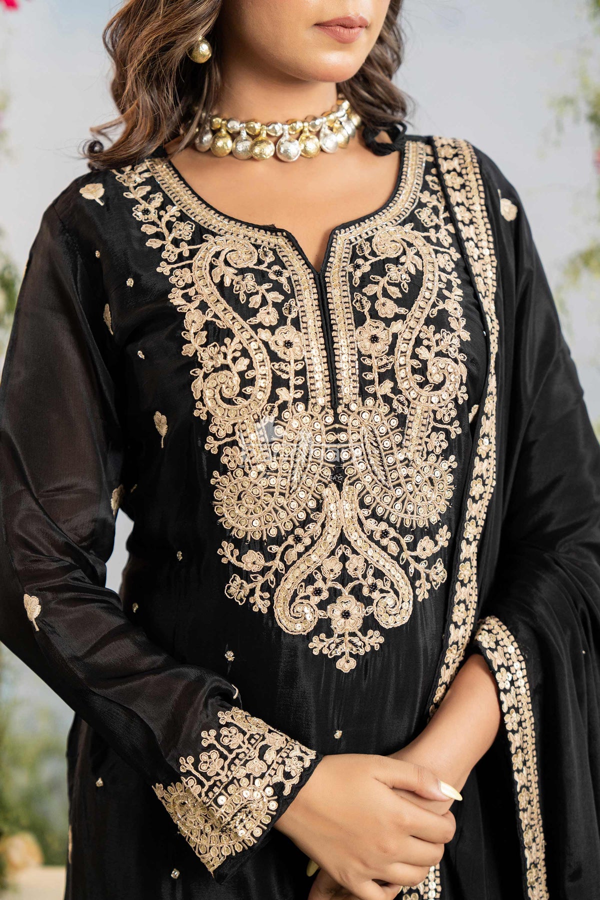 Black traditional outfit with gold embroidery and a pearl necklace against a blurred natural background