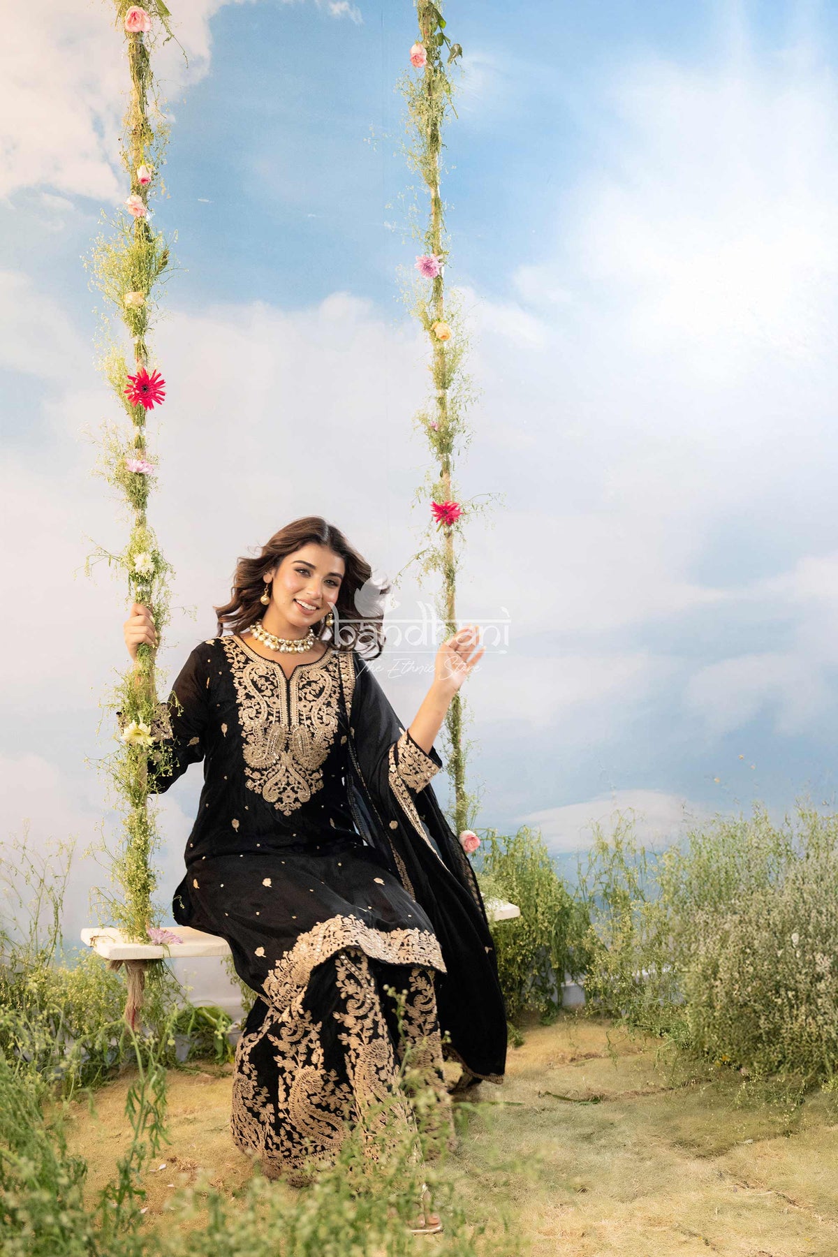 Woman in traditional black and gold outfit on a swing with floral decorations against a blue sky.