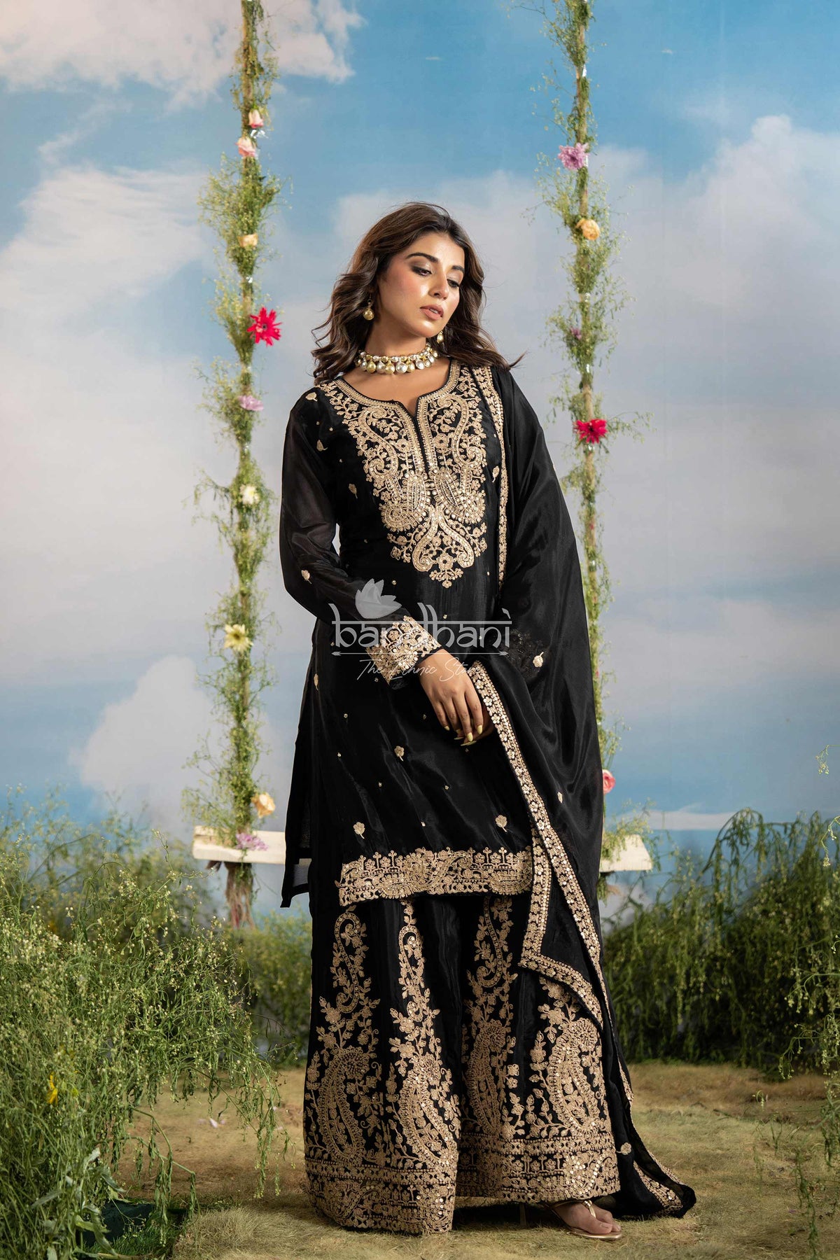 Woman in black embroidered traditional outfit standing on a swing with floral decorations against a blue sky.