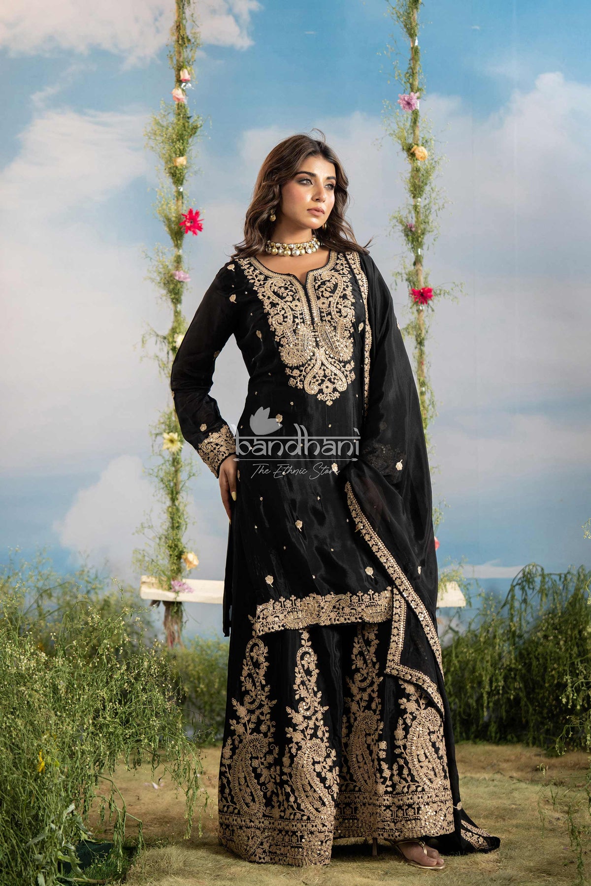 Woman in a black and gold embroidered traditional outfit standing outdoors with floral decorations.