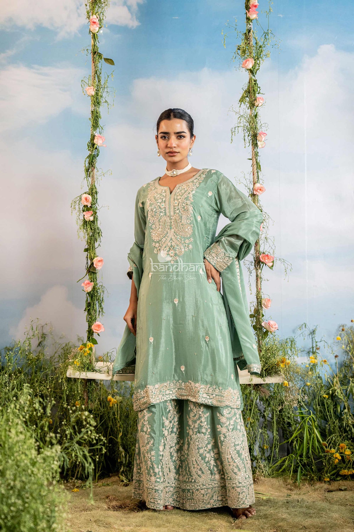Woman in a light green traditional outfit standing on a swing with floral decorations against a blue sky.