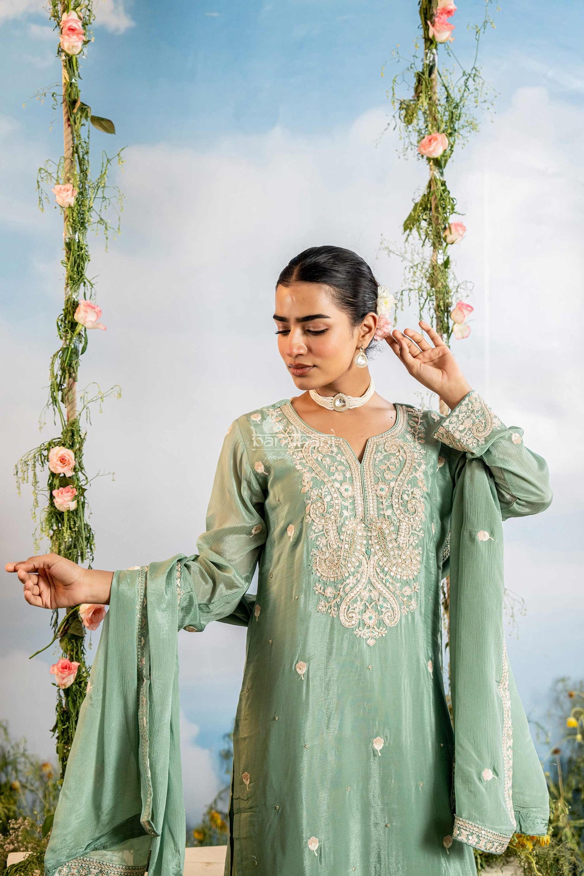 Woman in a light green embroidered traditional outfit standing outdoors with floral decorations.