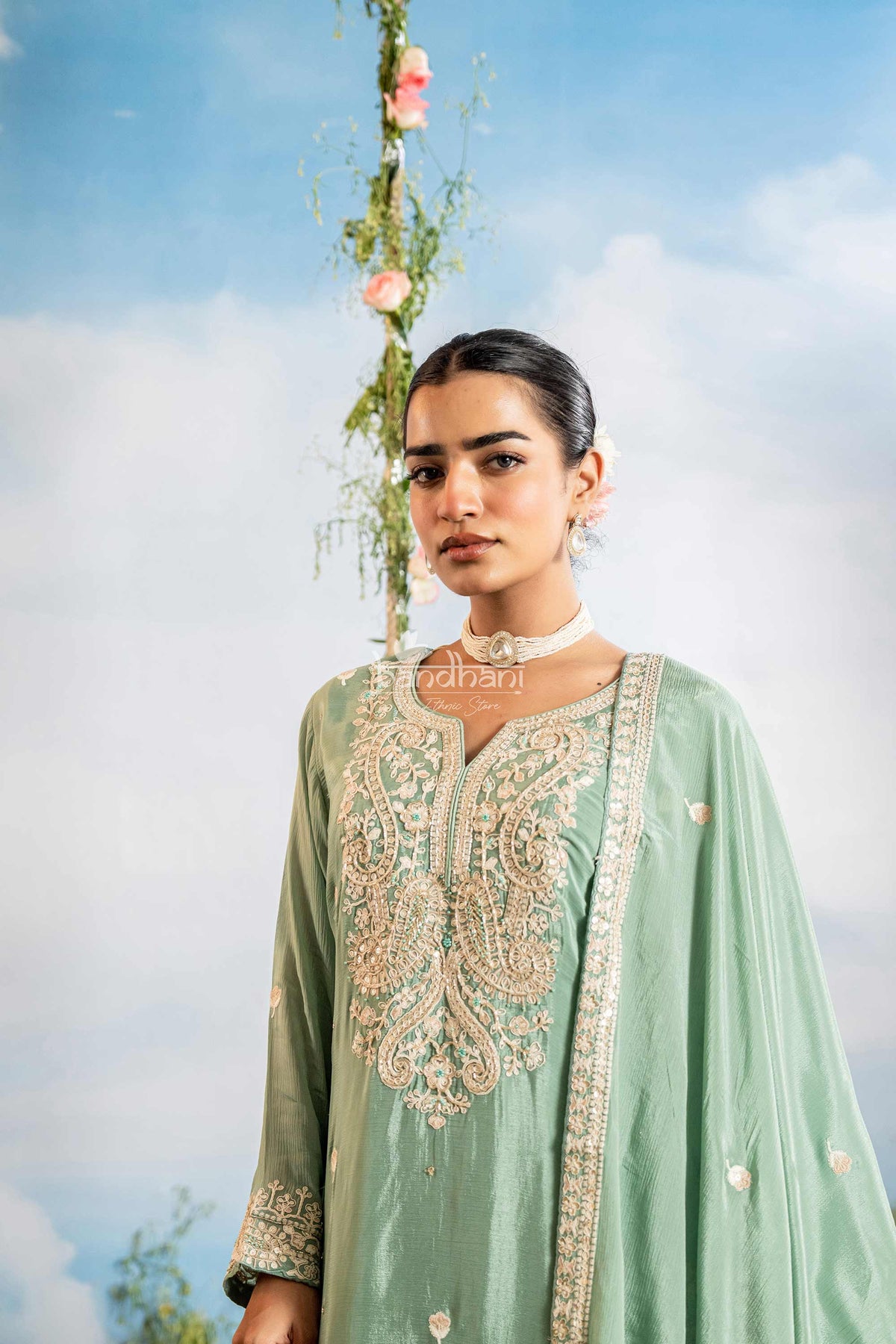 Woman wearing a light green embroidered traditional outfit against a blue sky with flowers.