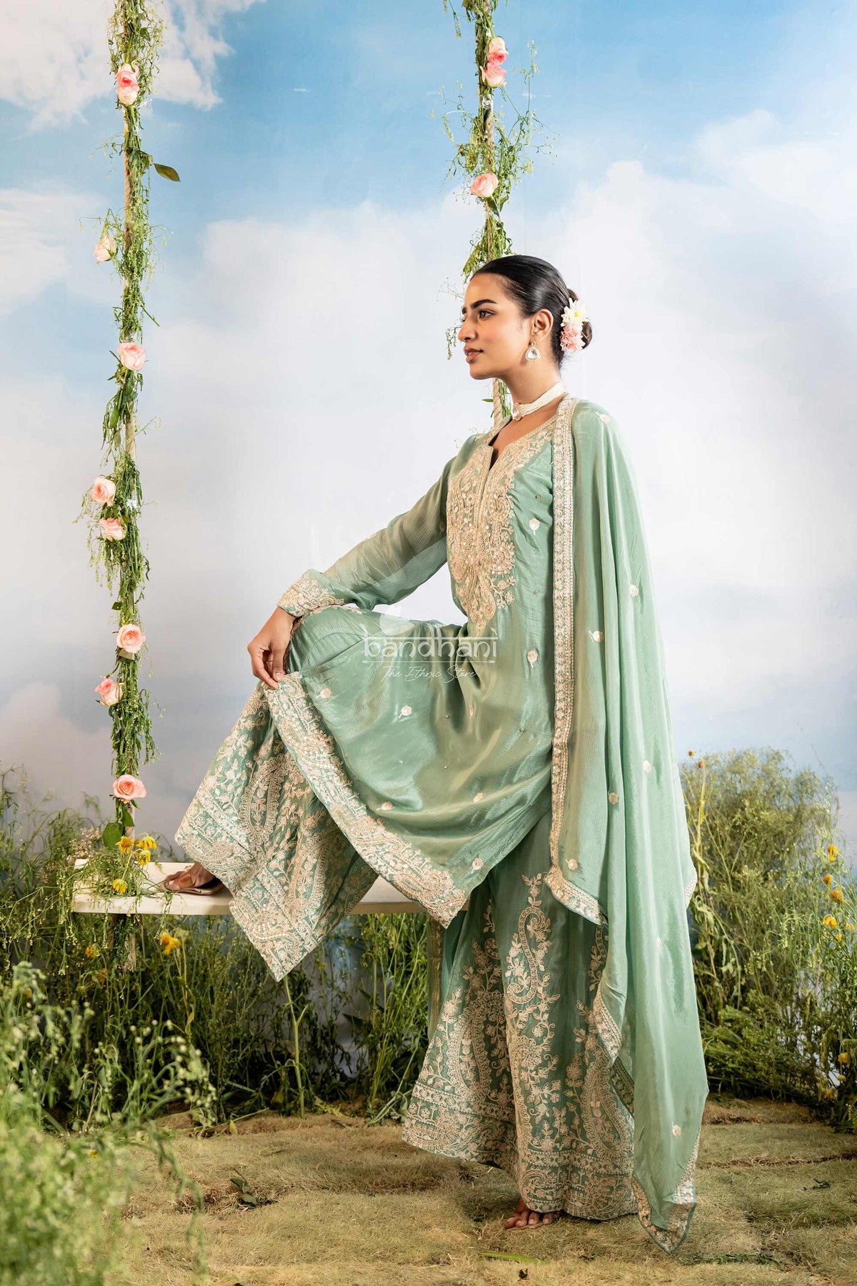 Woman in a light green traditional outfit sitting on a swing with floral decorations against a blue sky.