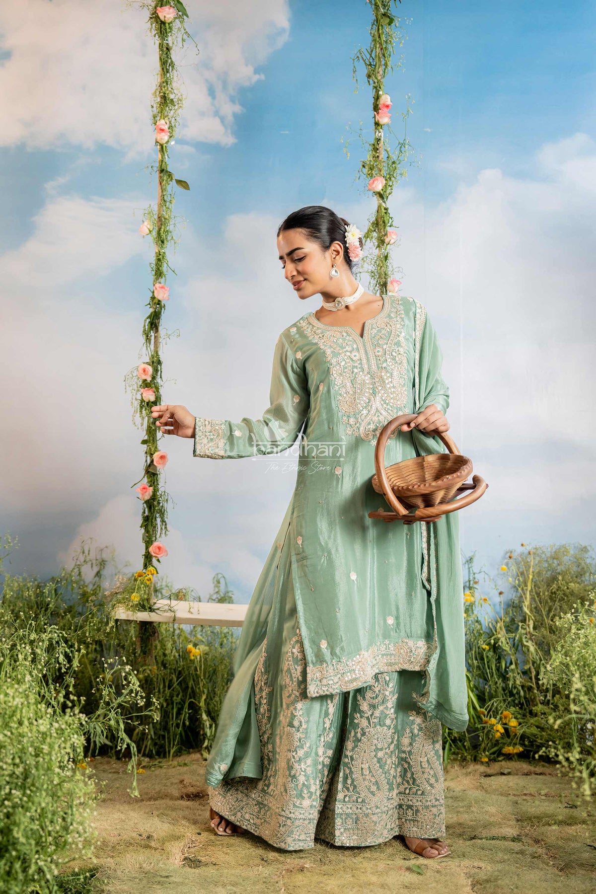 Woman in a green traditional outfit standing next to a swing with floral decorations against a blue sky.