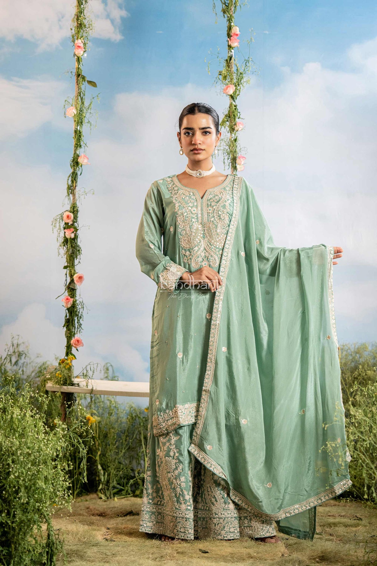 Woman in a light green traditional outfit with floral decorations against a blue sky.