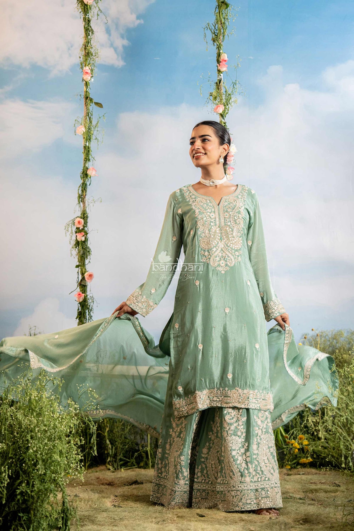 Woman in a light green traditional outfit with floral decorations against a blue sky.