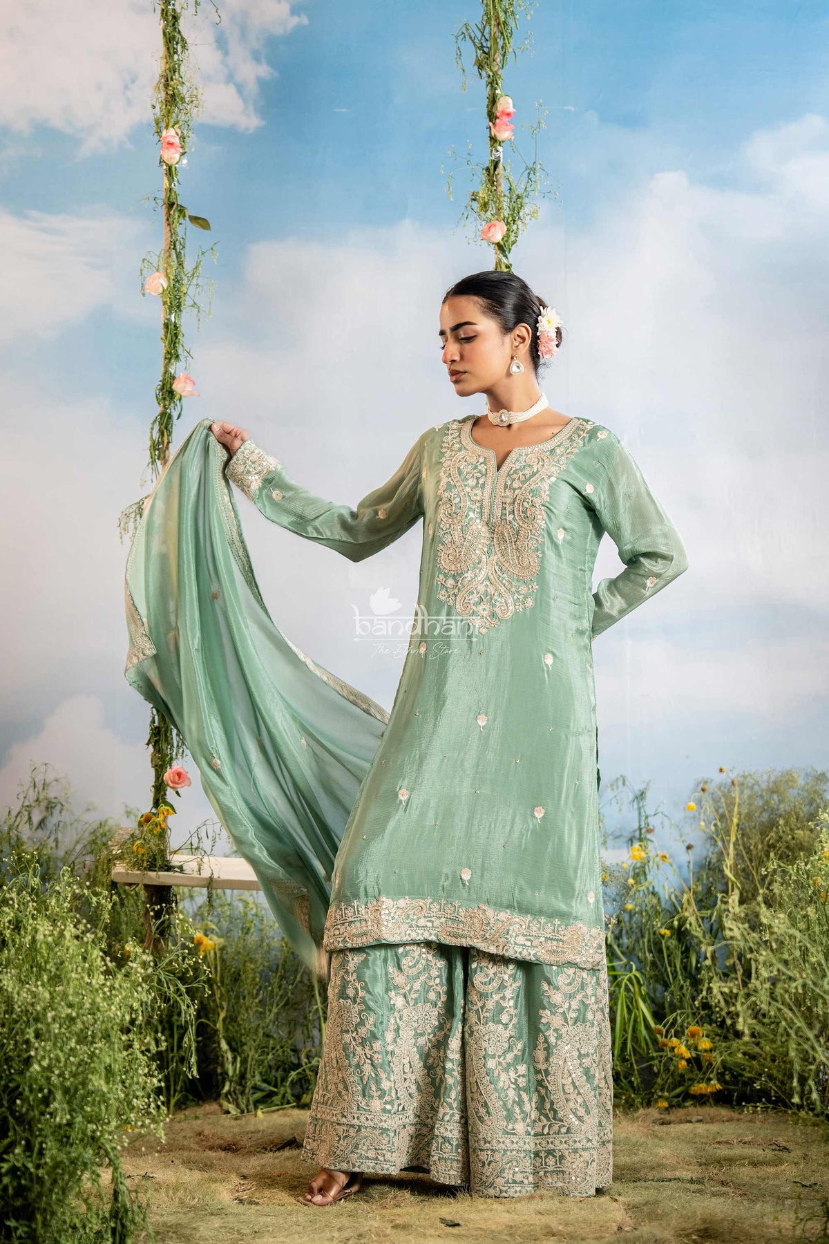 Woman in a light green traditional outfit with floral patterns standing outdoors against a blue sky.