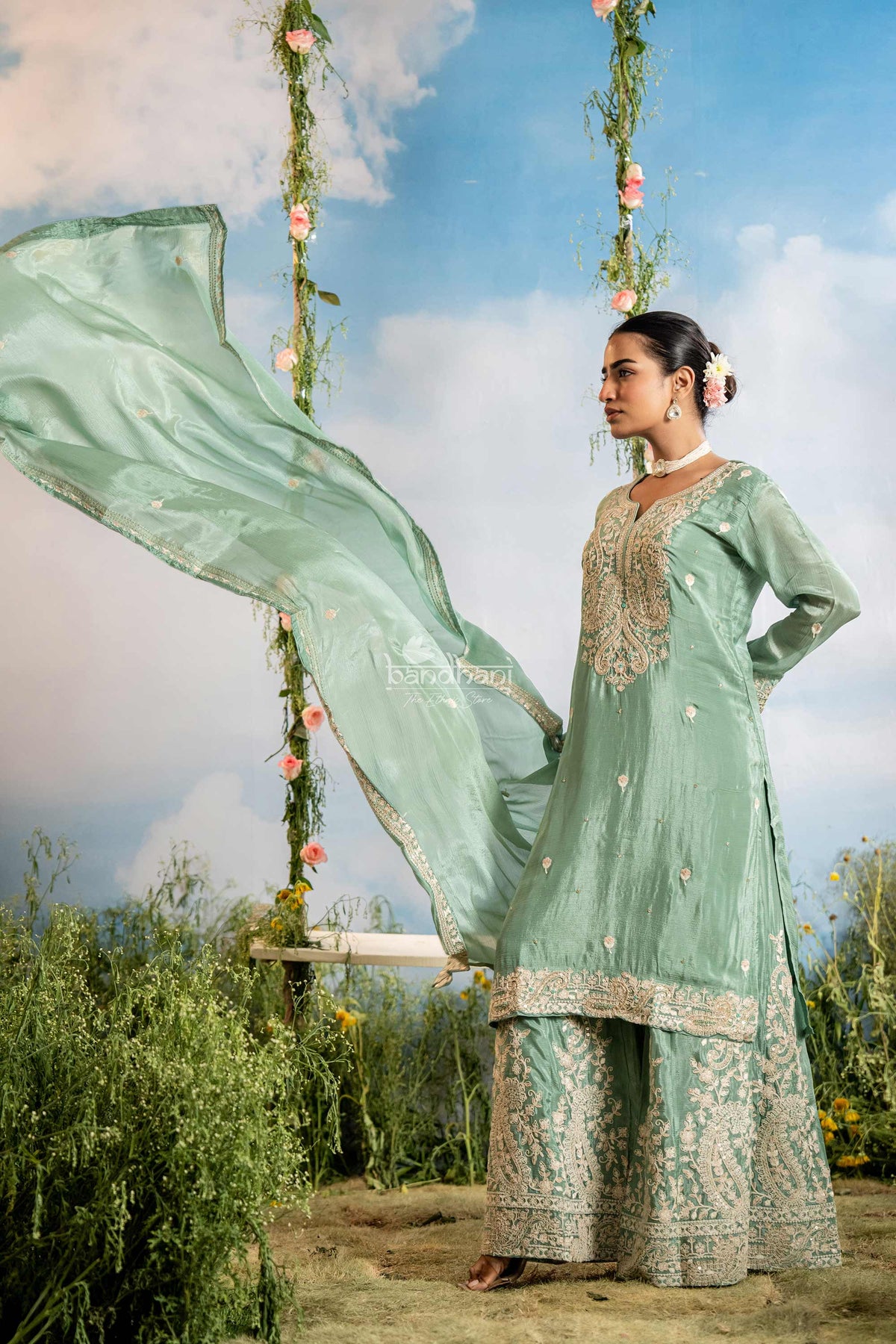Woman in a light green traditional outfit with floral decorations against a blue sky.
