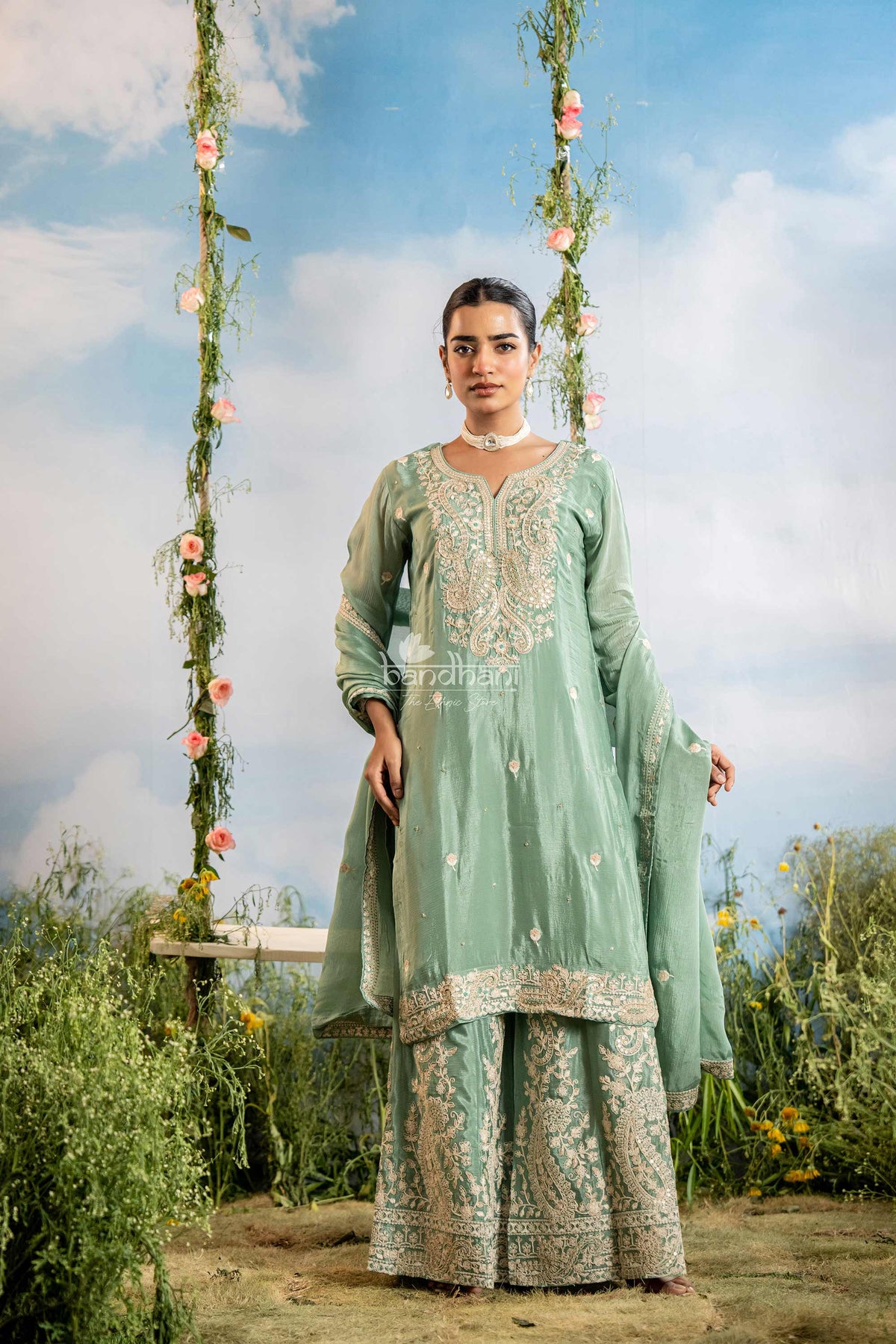 Woman in a traditional outfit standing on a swing with floral decorations against a blue sky.