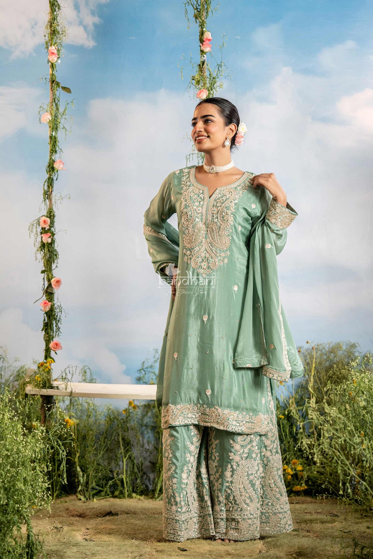 Woman in a traditional outfit standing outdoors with a floral swing and blue sky in the background