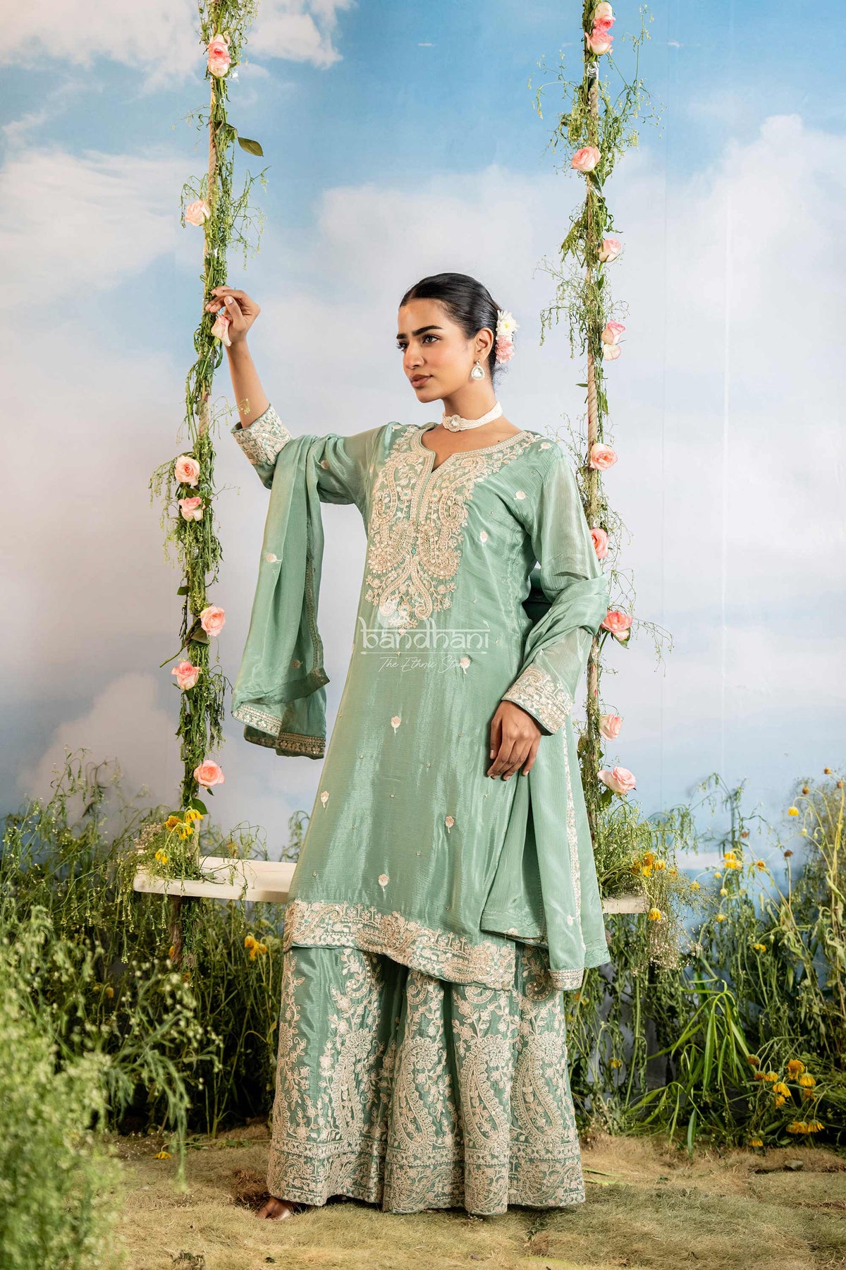 Woman in a light green traditional outfit sitting on a swing with floral decorations against a blue sky.