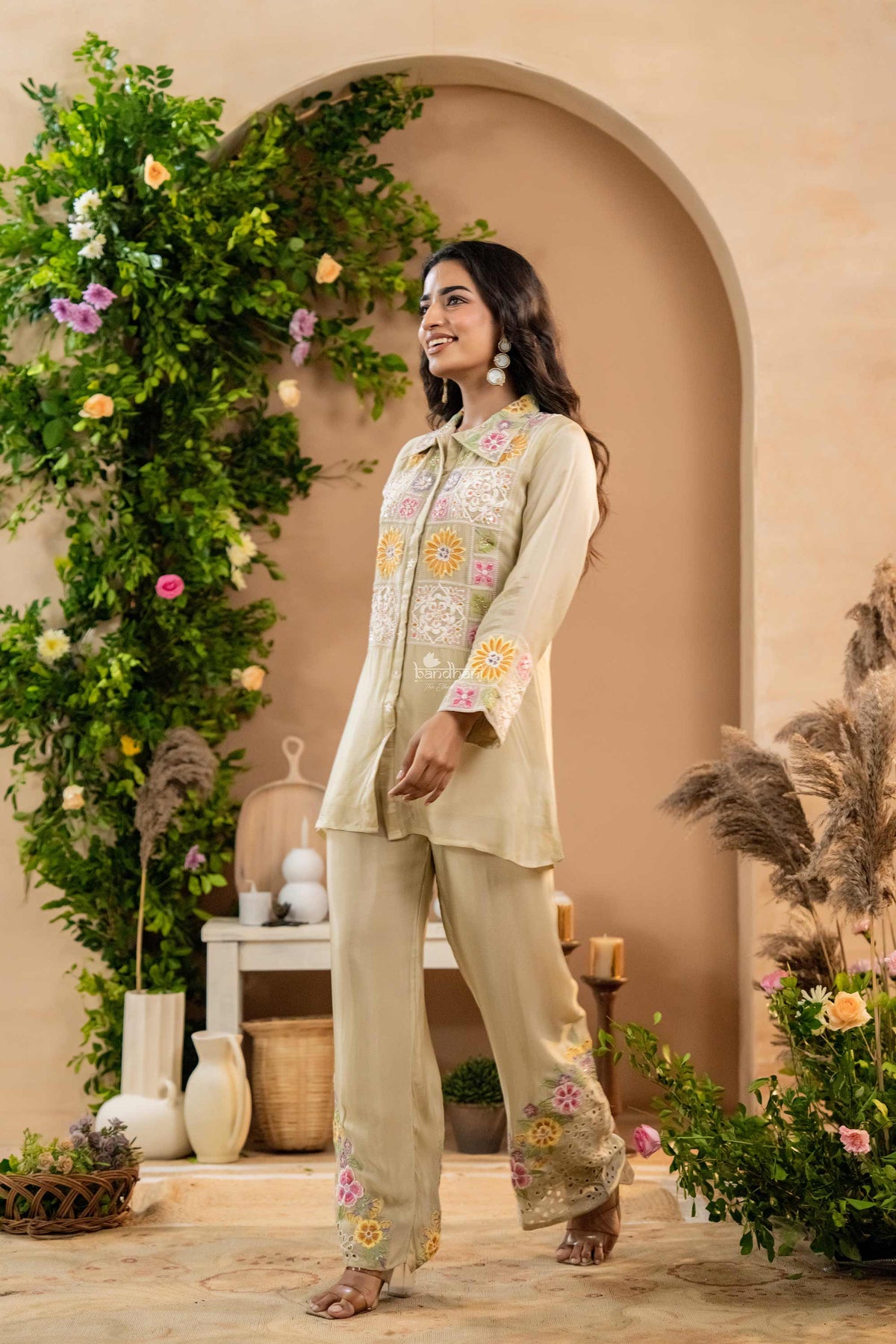 Woman in a floral embroidered outfit standing in a decorated indoor setting with plants and archway.