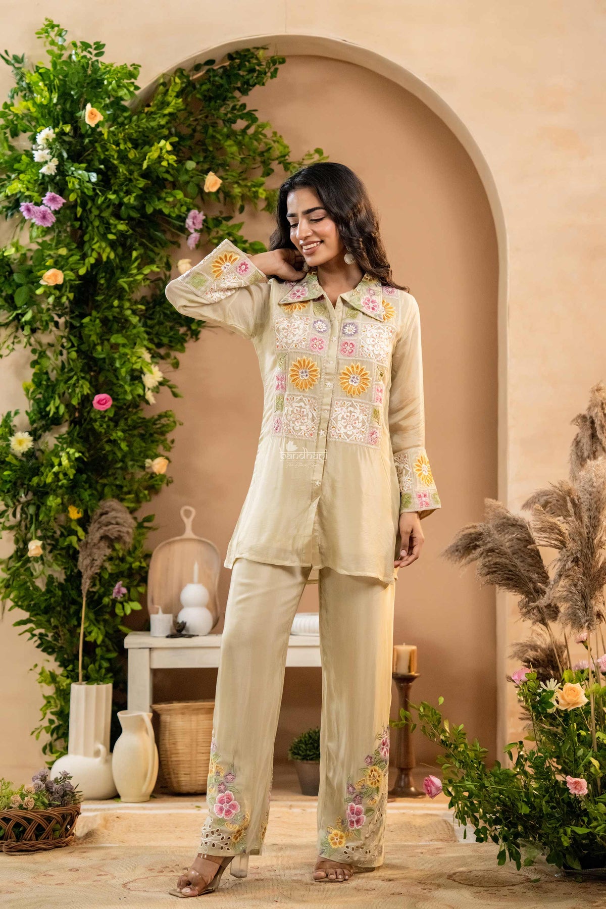 Woman in a cream embroidered outfit standing in a decorated indoor setting with plants and decor.