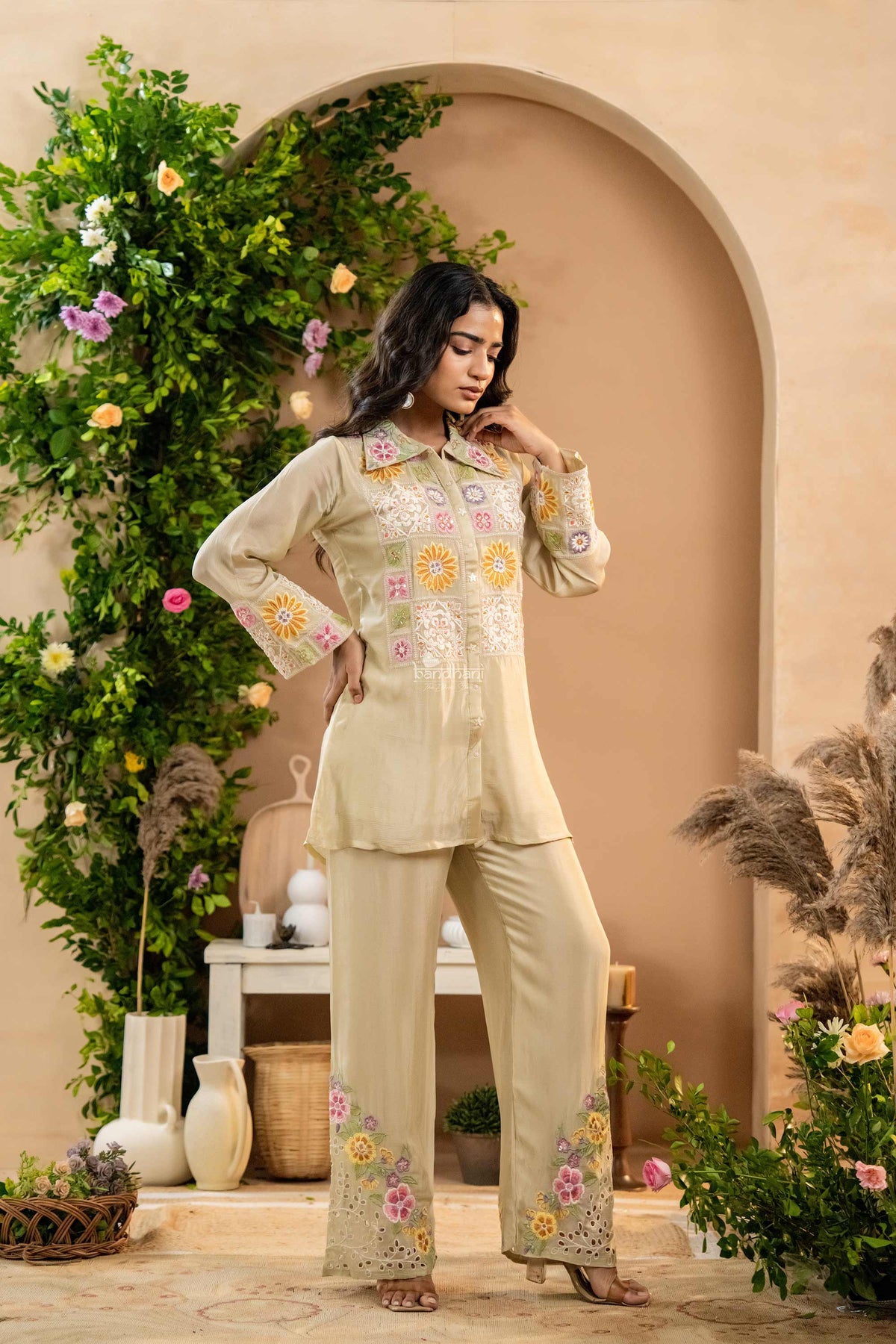 Woman in a light green embroidered outfit standing in a decorated outdoor setting with plants and flowers.