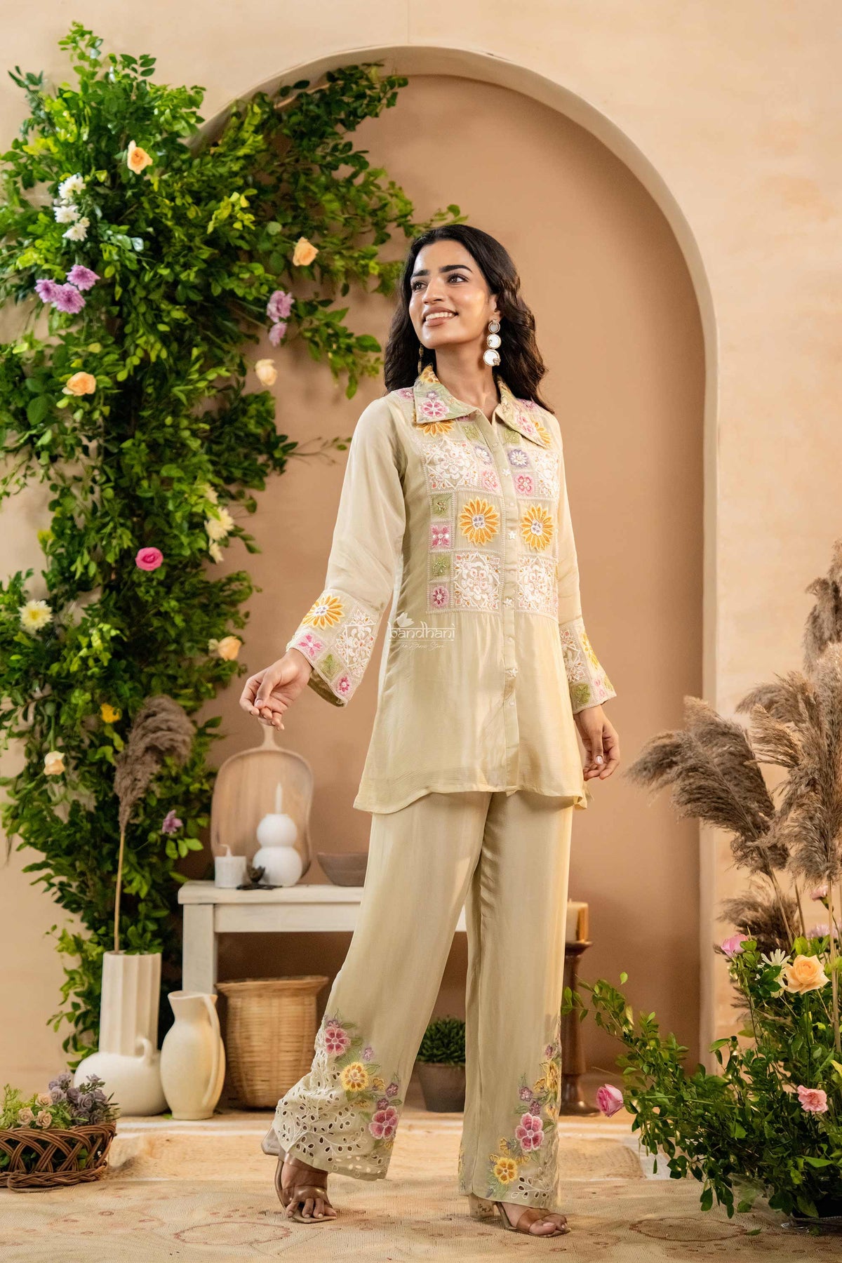 Woman in a traditional outfit standing in a decorated room with plants and flowers.