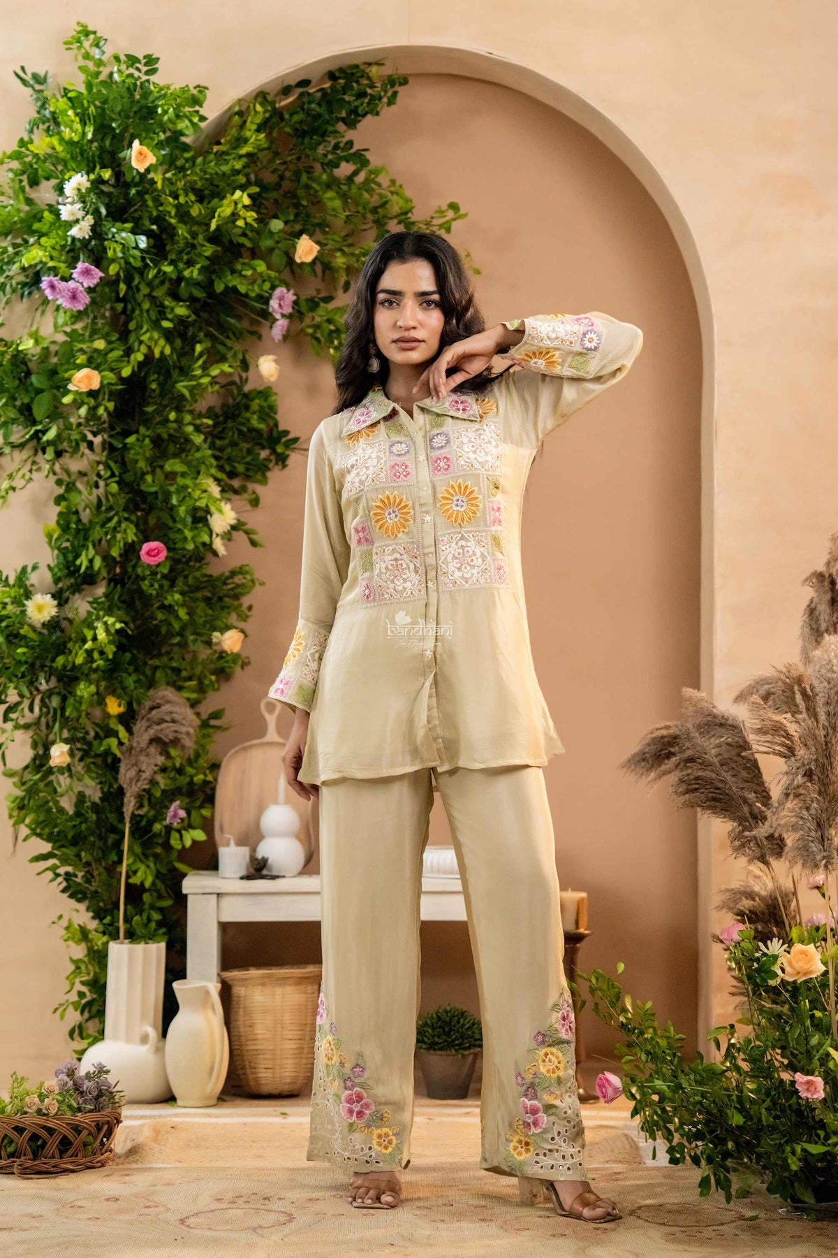 Woman in a light green embroidered outfit standing in a decorated indoor setting with plants and flowers.