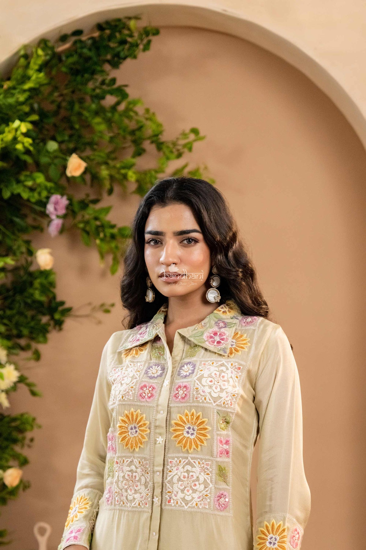 Woman wearing a light-colored embroidered traditional outfit with a beige wall and greenery in the background