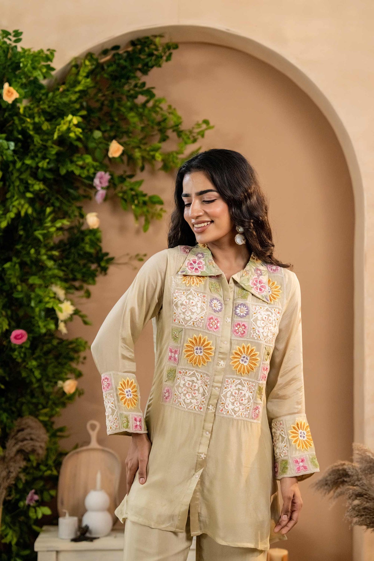 Woman wearing a beige embroidered co-ord set standing in front of a decorative archway with greenery.