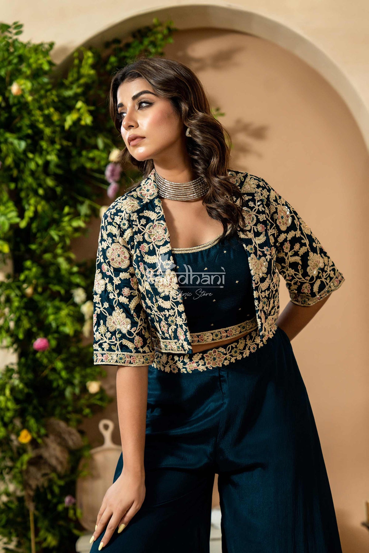 Woman wearing a floral blouse over a black top with visible branding, standing against a beige wall with greenery.