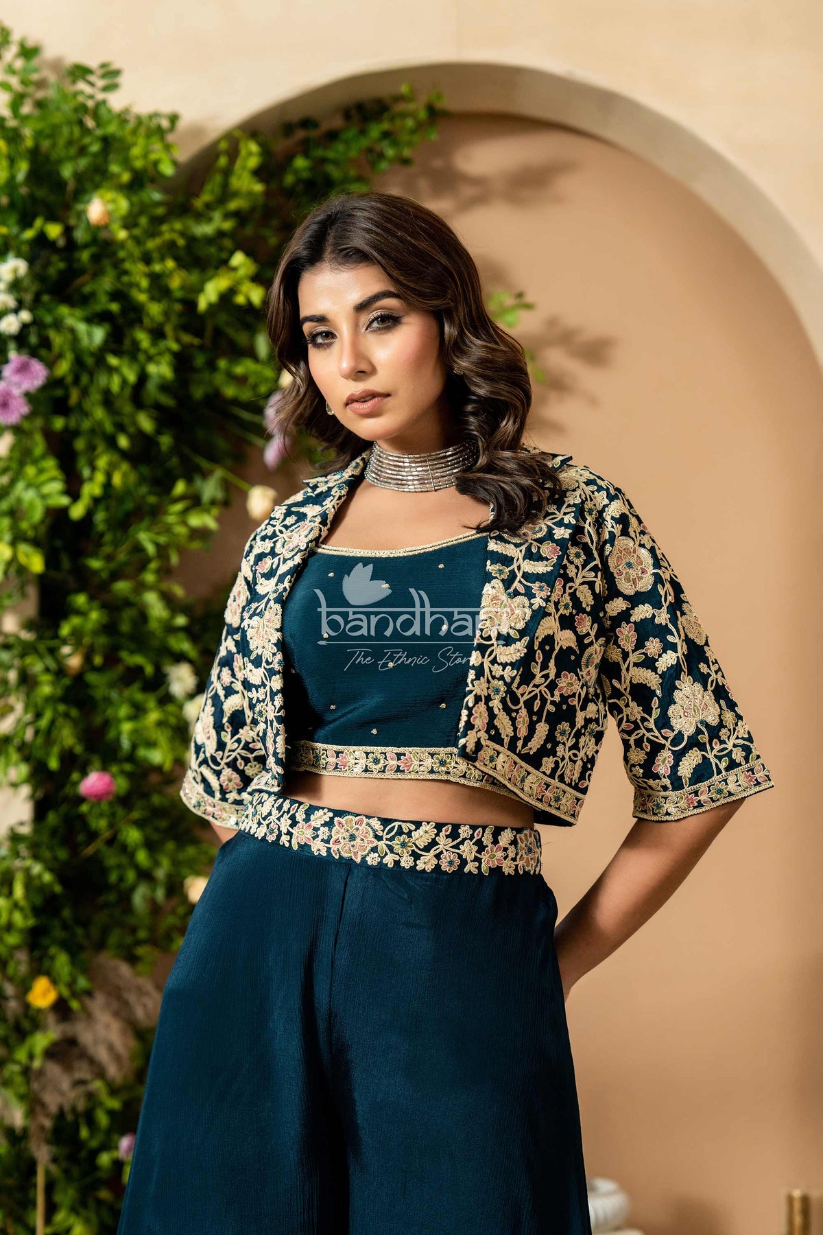 Woman wearing a dark blue traditional outfit with floral patterns, standing against a beige wall with greenery.