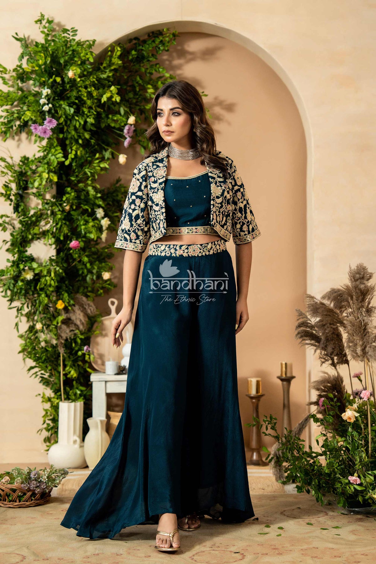 Woman in a blue outfit with a decorative jacket standing in front of an archway with greenery.