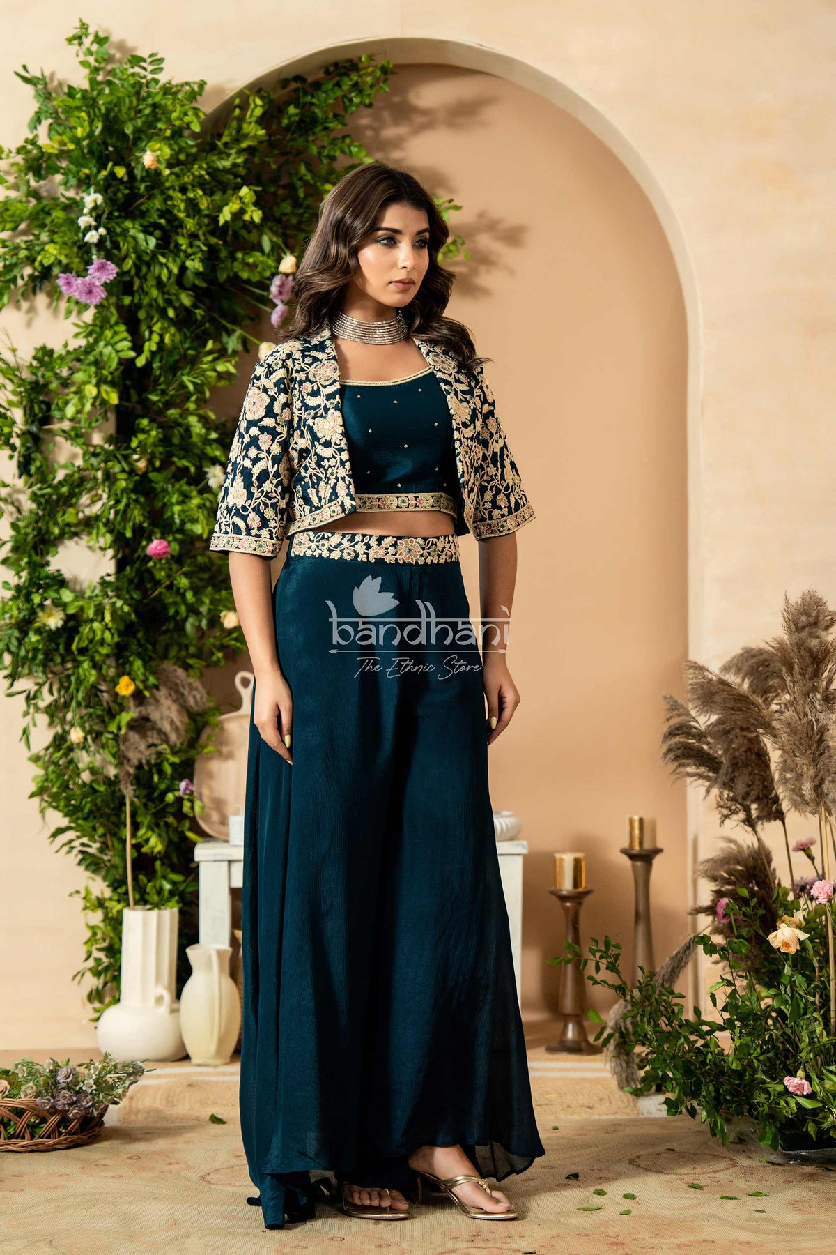Woman in a teal outfit with a floral jacket standing in front of an archway with decorative plants.