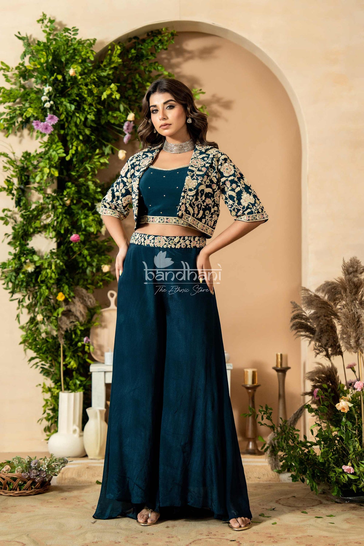 Woman in a blue traditional outfit with floral decorations, standing in front of an archway with greenery.