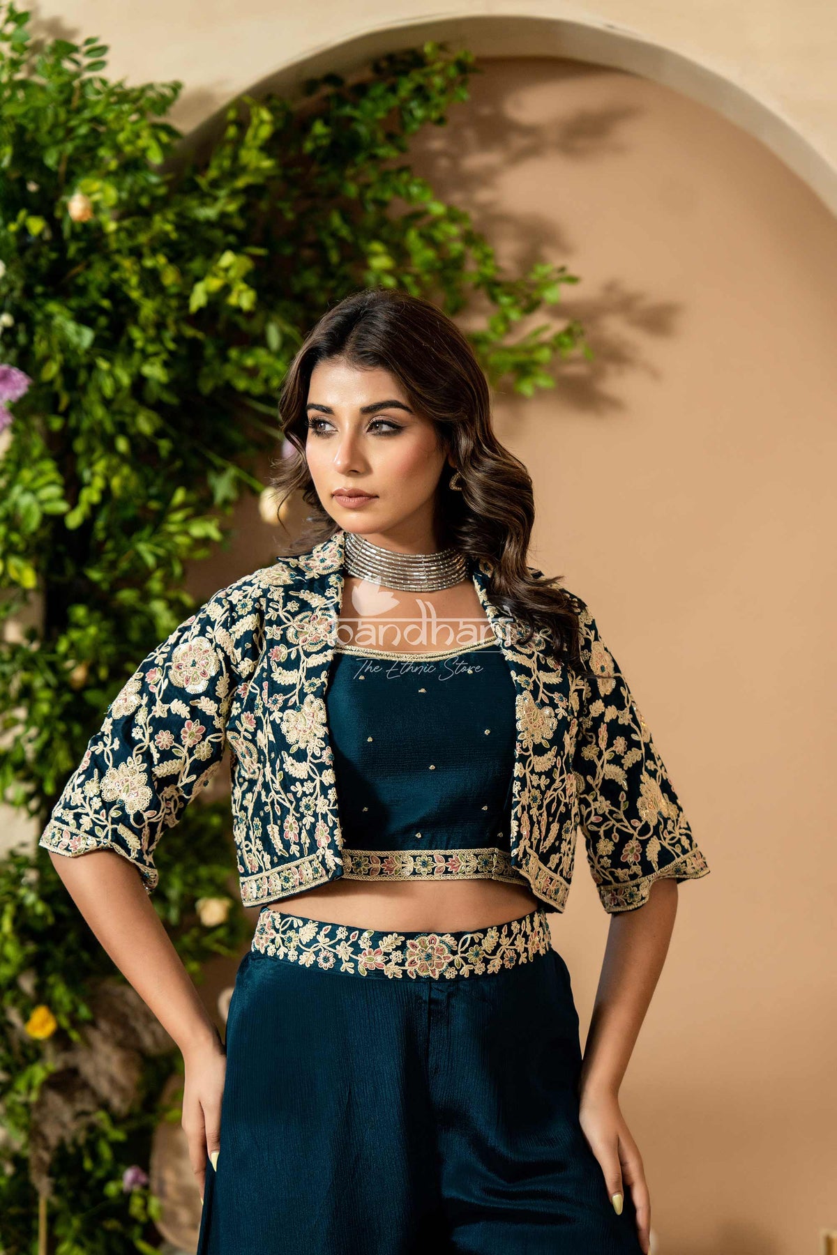 Woman wearing a traditional outfit with a floral jacket and dark blue saree against a beige wall.