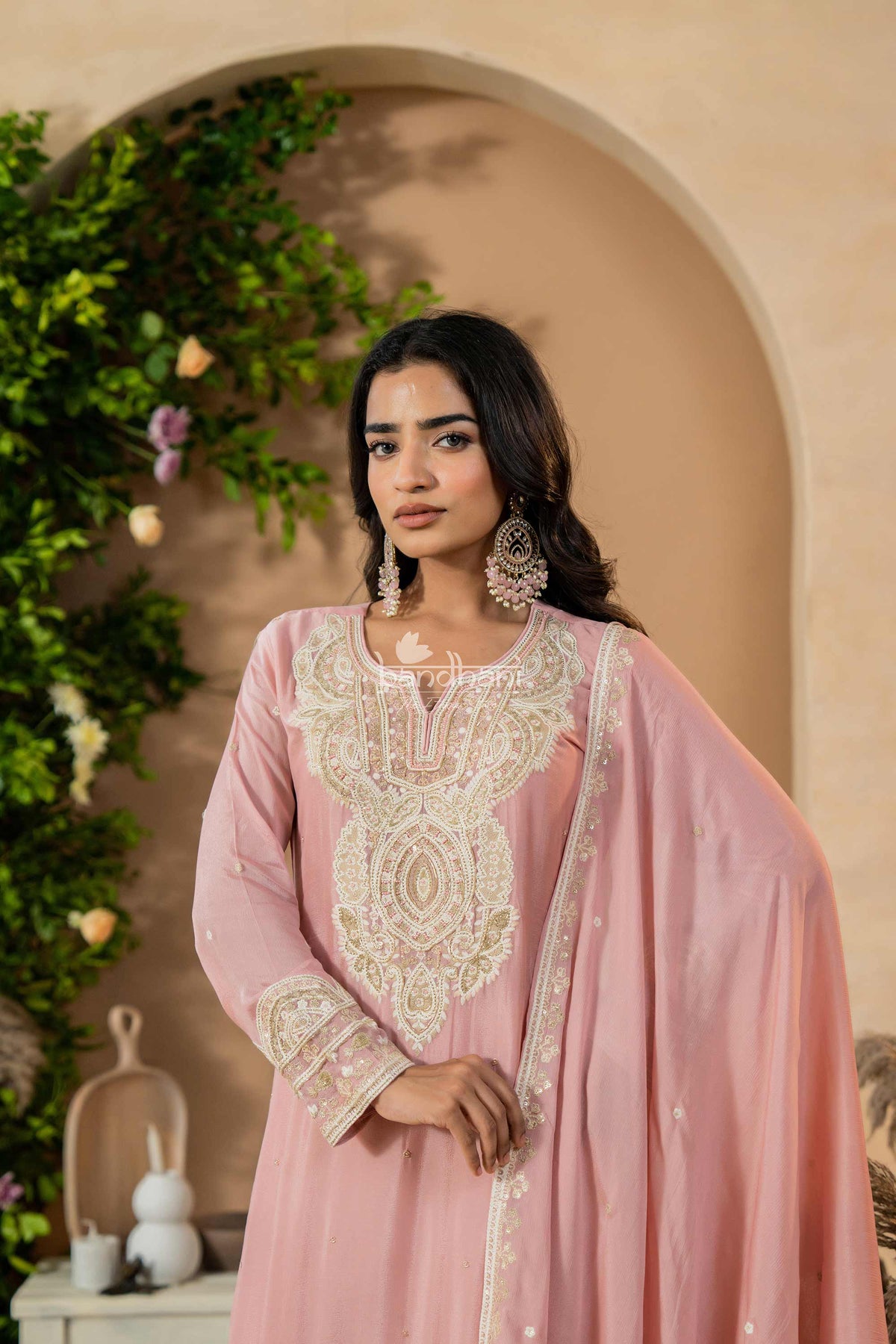 Woman wearing a pink embroidered traditional kurti pant dupatta set with a beige wall and greenery in the background