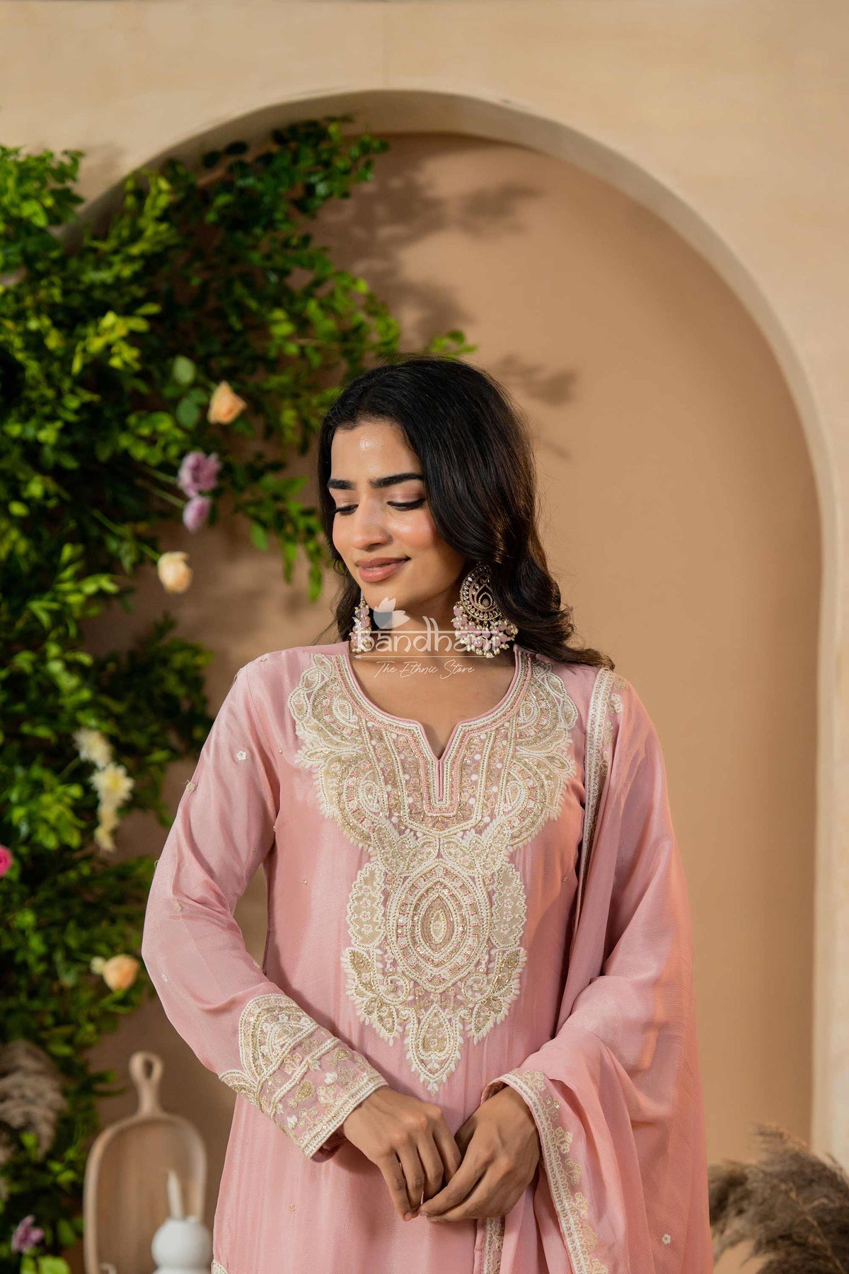 Woman in a pink traditional outfit with intricate white embroidery, standing against a decorative background.