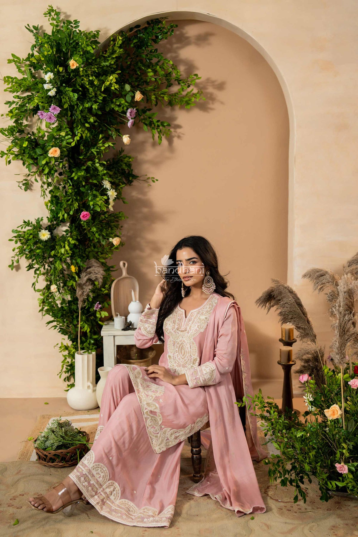 Woman in a pink traditional outfit sitting in a decorated room with plants and flowers.