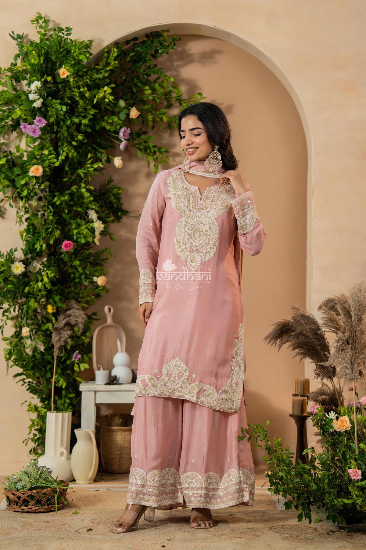 Woman in a pink traditional outfit with white embroidery standing in a decorated indoor setting.
