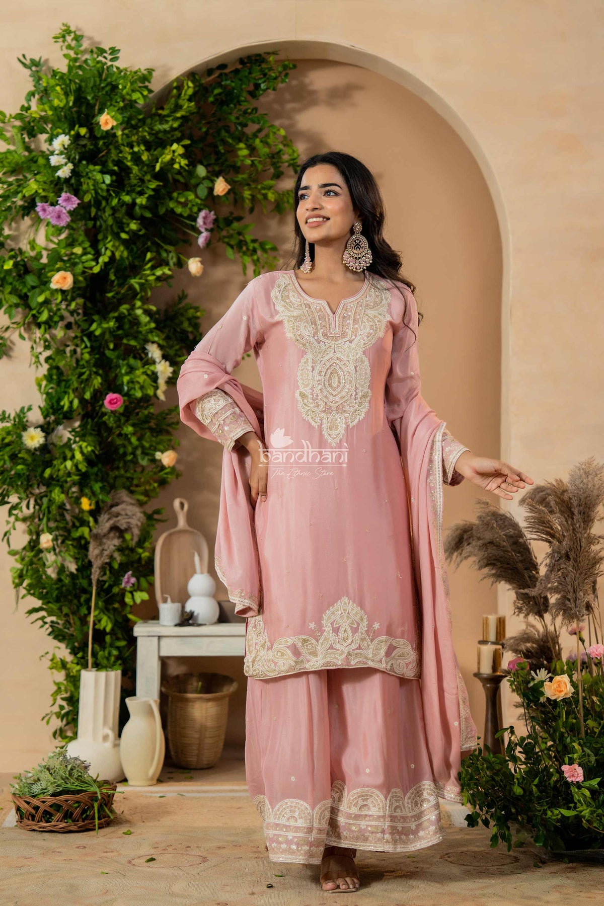 Woman in a pink traditional outfit with intricate embroidery standing in a decorated indoor setting.