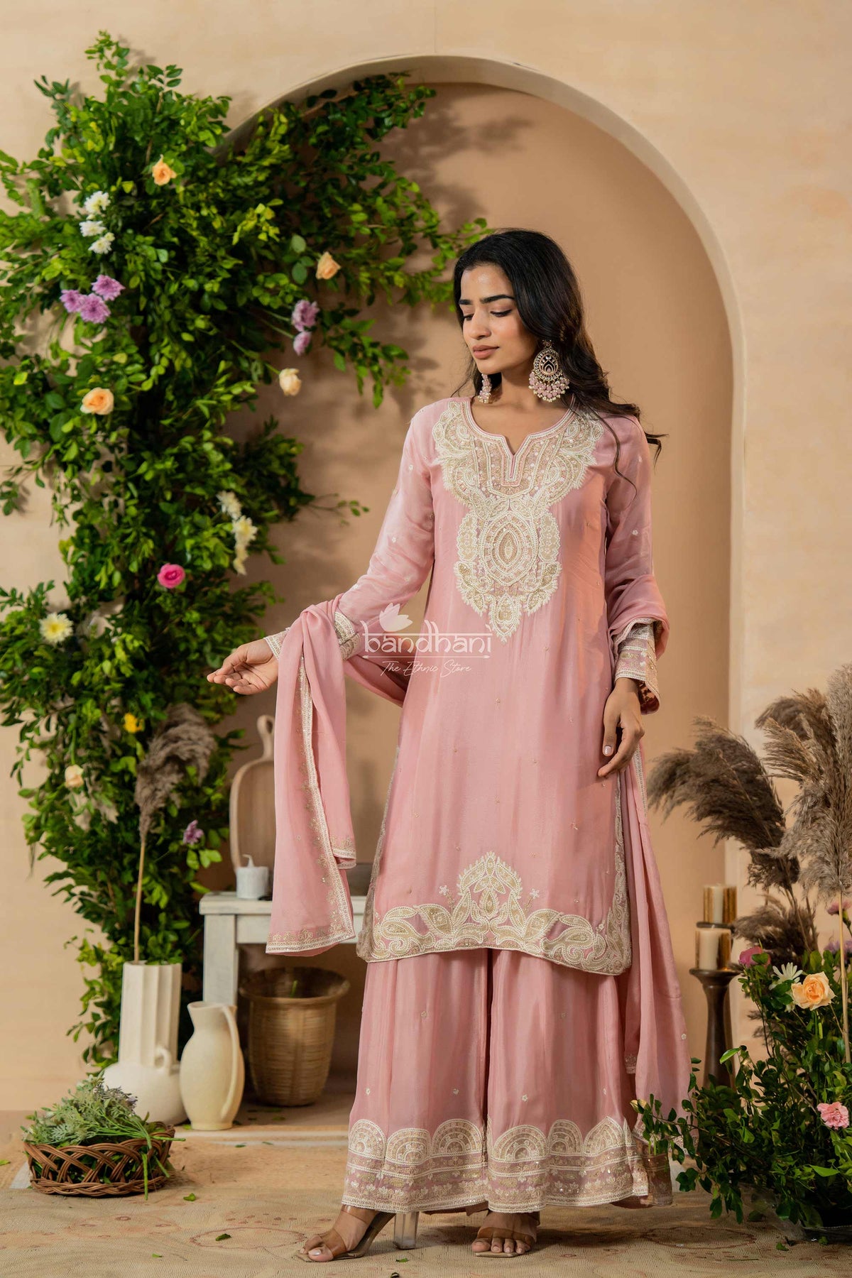 Woman in a pink traditional outfit with intricate white embroidery standing in a decorated indoor setting.