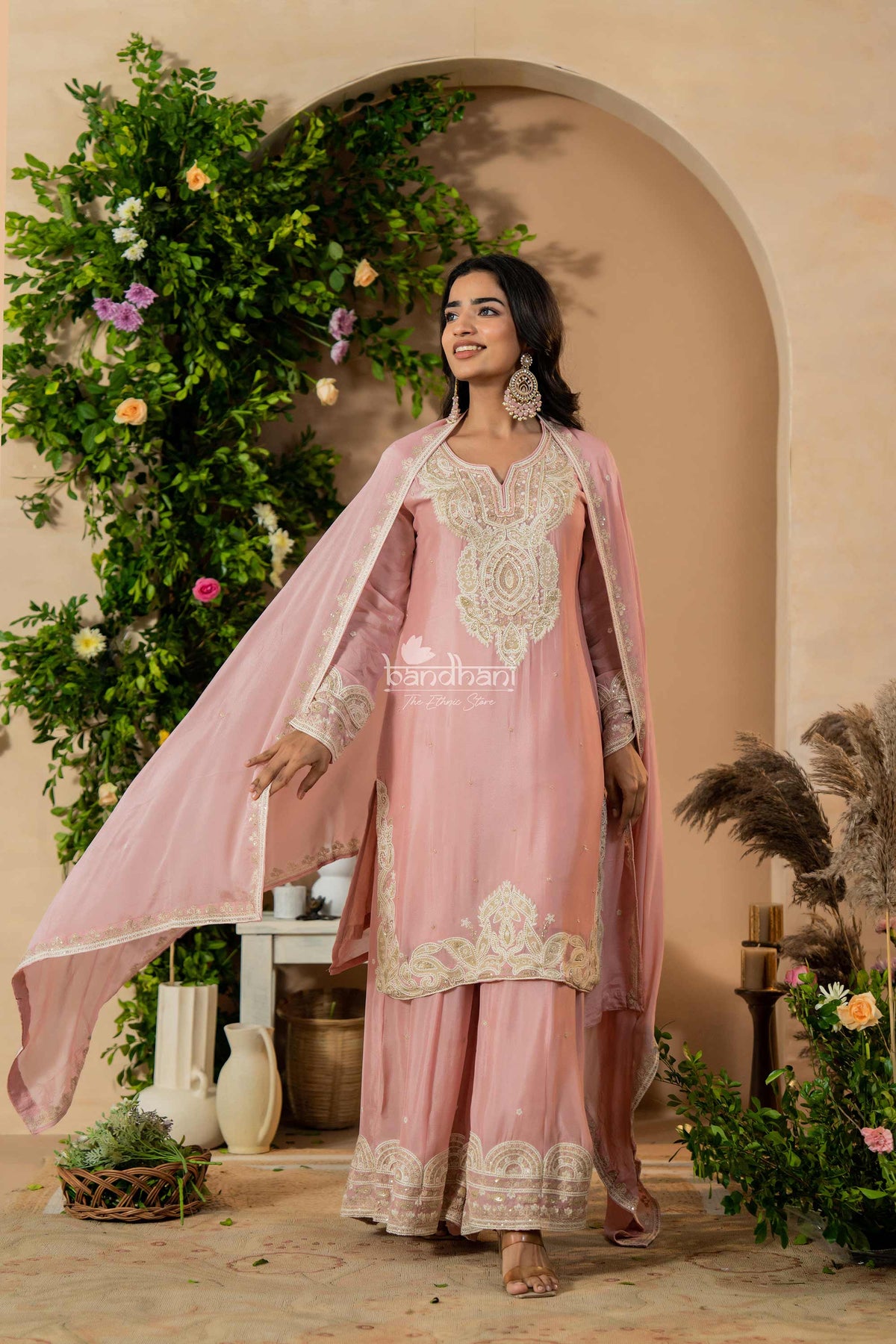 Woman in a pink traditional outfit with white embroidery standing in a decorated indoor setting.