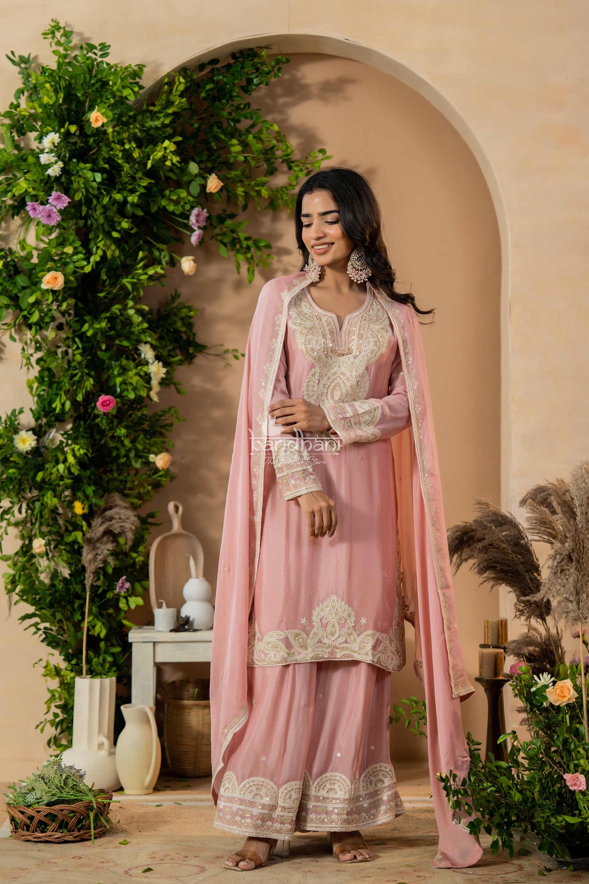 Woman in a pink traditional outfit standing in a decorated indoor setting with greenery and flowers.