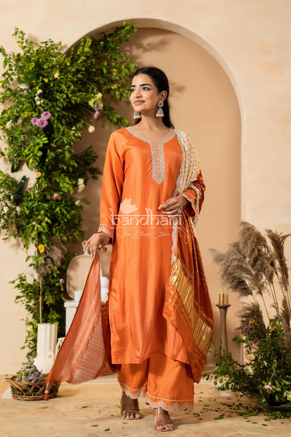 Woman in an orange traditional outfit standing in a decorated outdoor setting with plants and flowers.