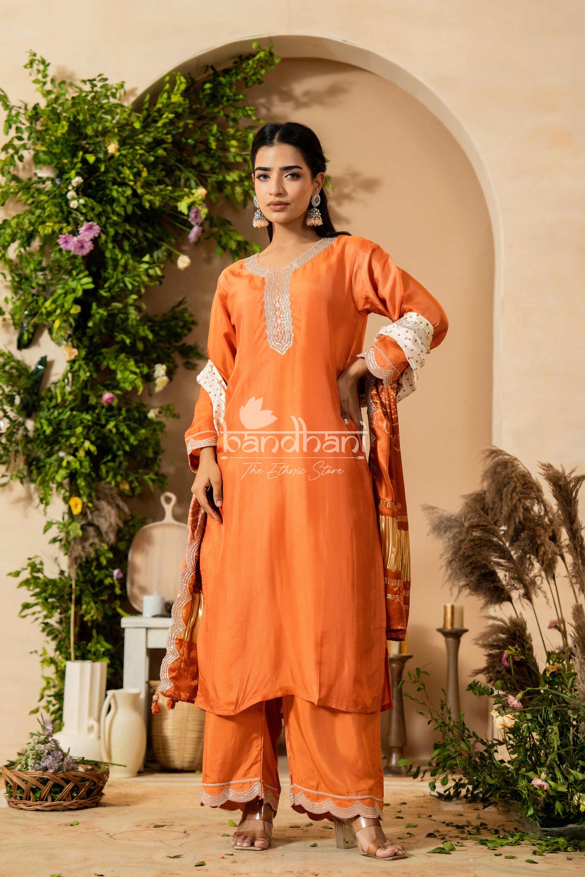 Woman in an orange traditional outfit standing in a decorated indoor setting with plants and archway.