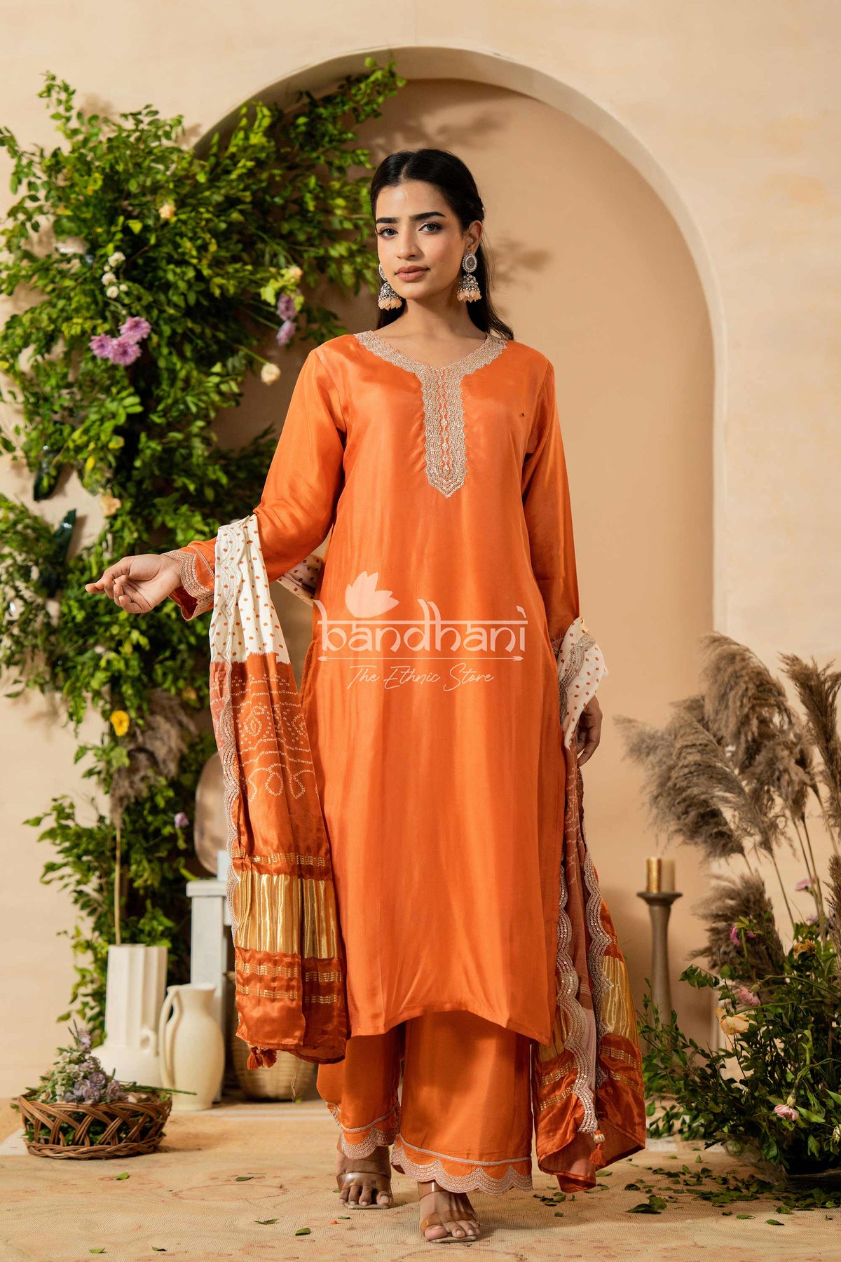 Woman wearing an orange traditional outfit with intricate designs, standing in a decorated outdoor setting.