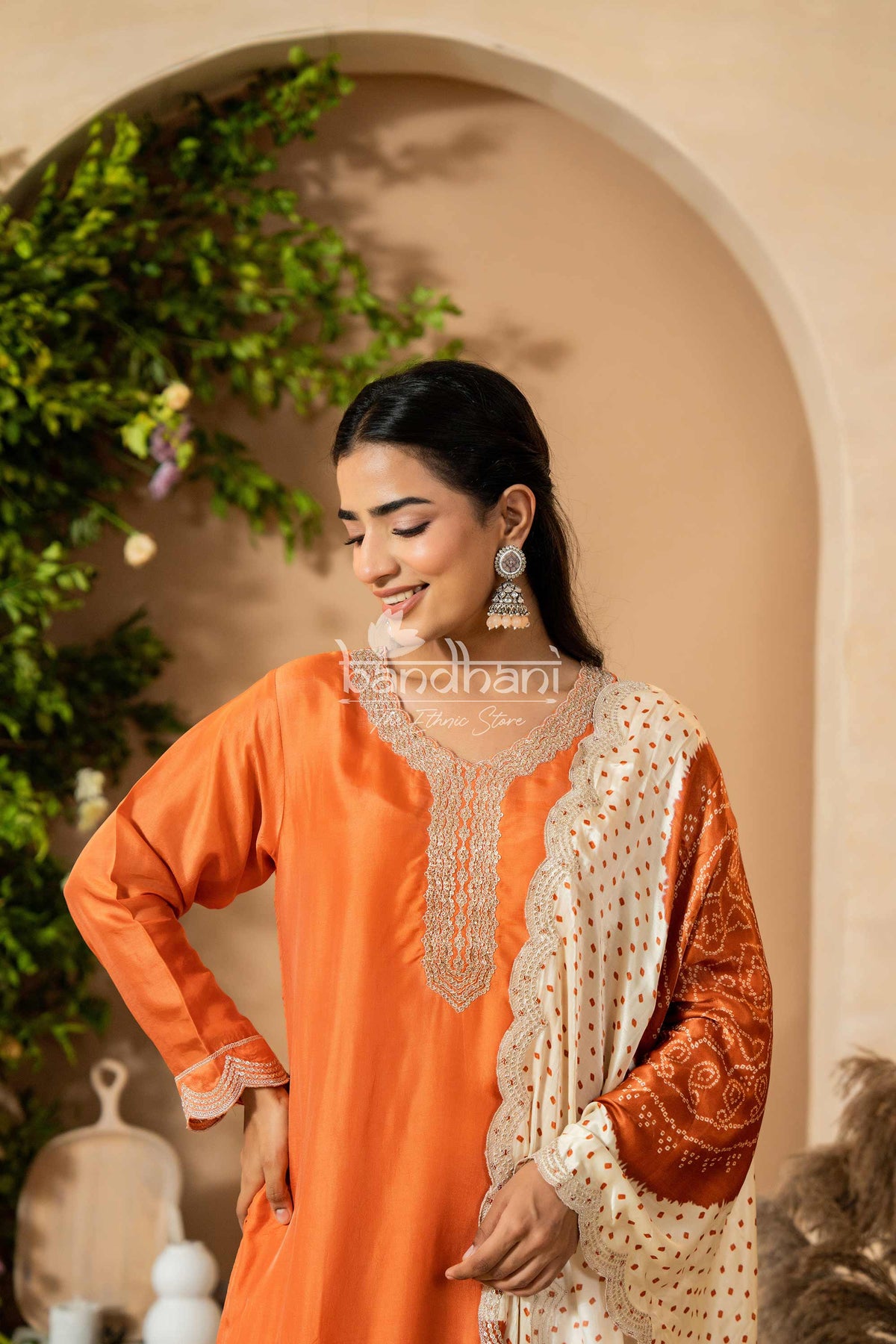 Woman wearing an orange and white traditional outfit with 'Bhandhani' branding, standing in a decorative setting.