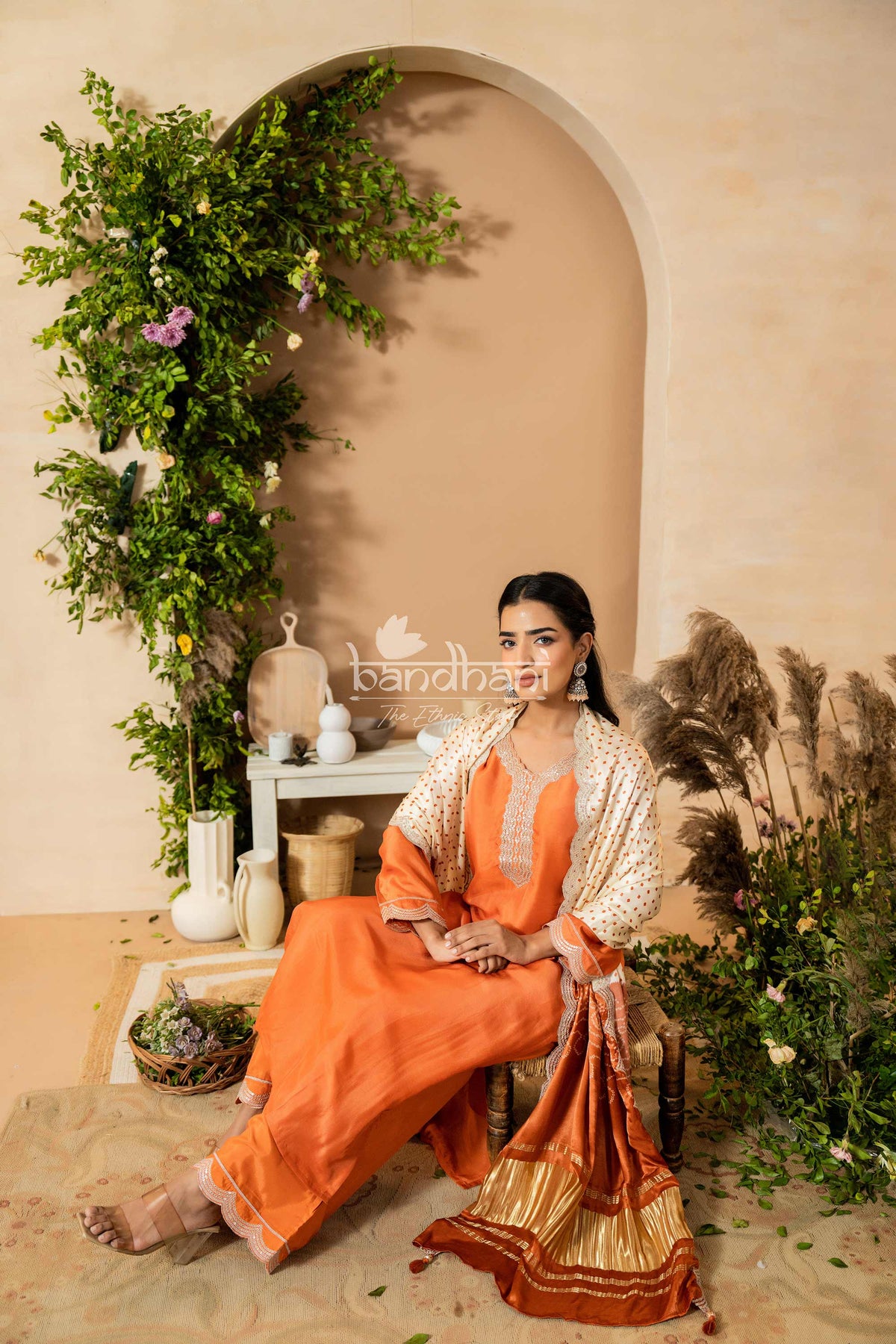 Woman in an orange and gold traditional outfit sitting in a decorated room with plants and decor.